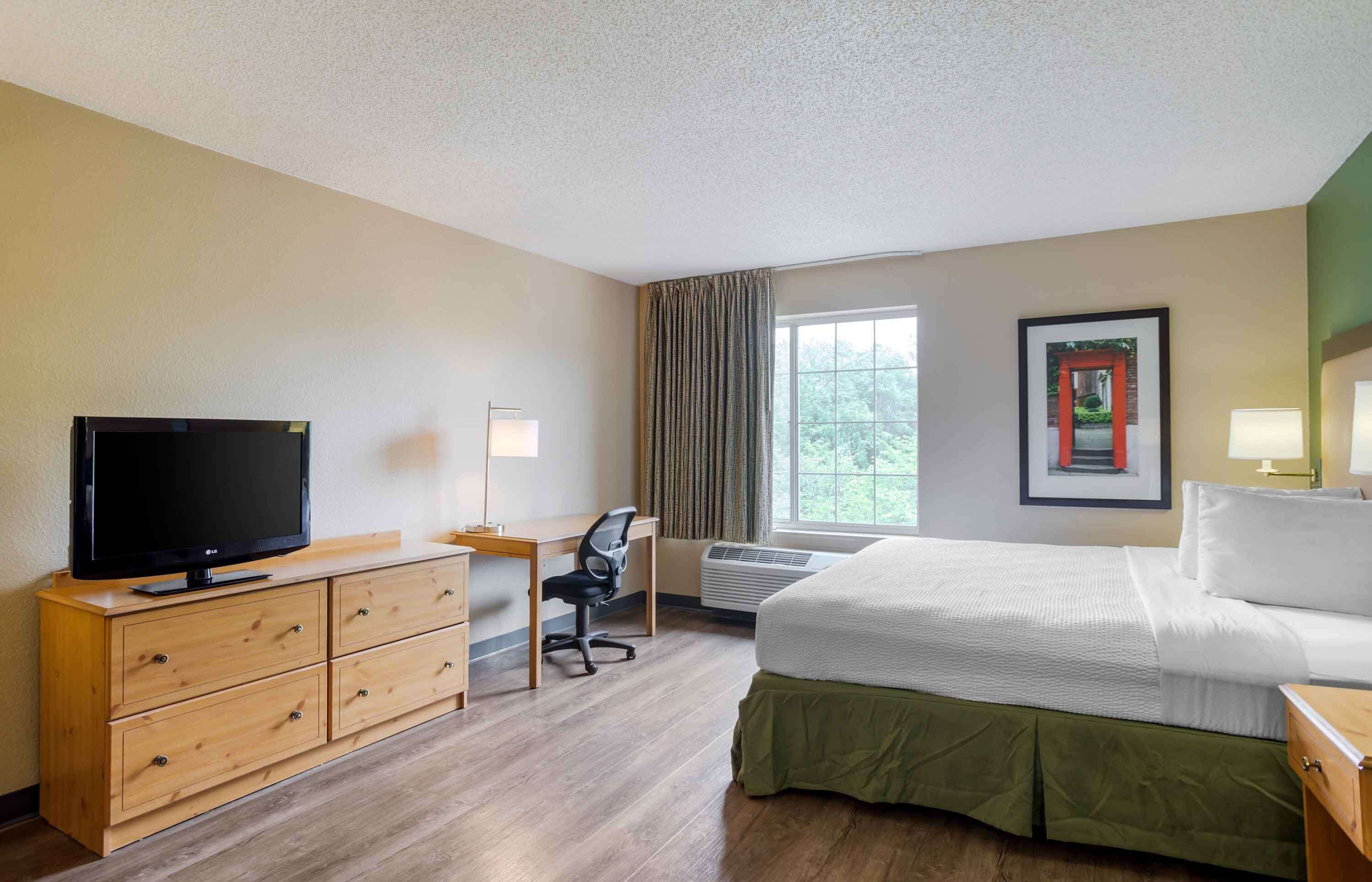 A hotel room with a bed, a desk with a chair, a television, and an air conditioning unit.