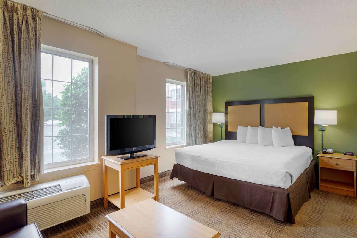 A hotel room with a large bed, a TV on a small table, a heater, and a nightstand with a lamp.