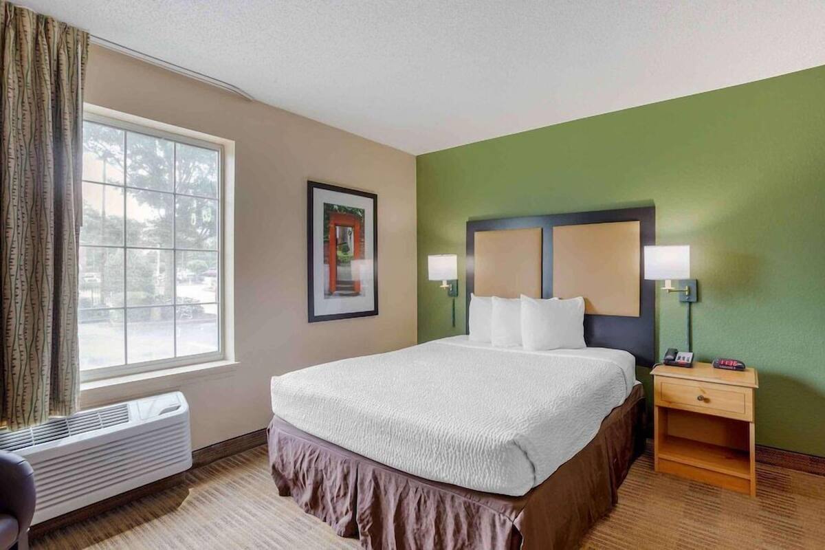 A hotel room with a bed, bedside table, lamp, and window.