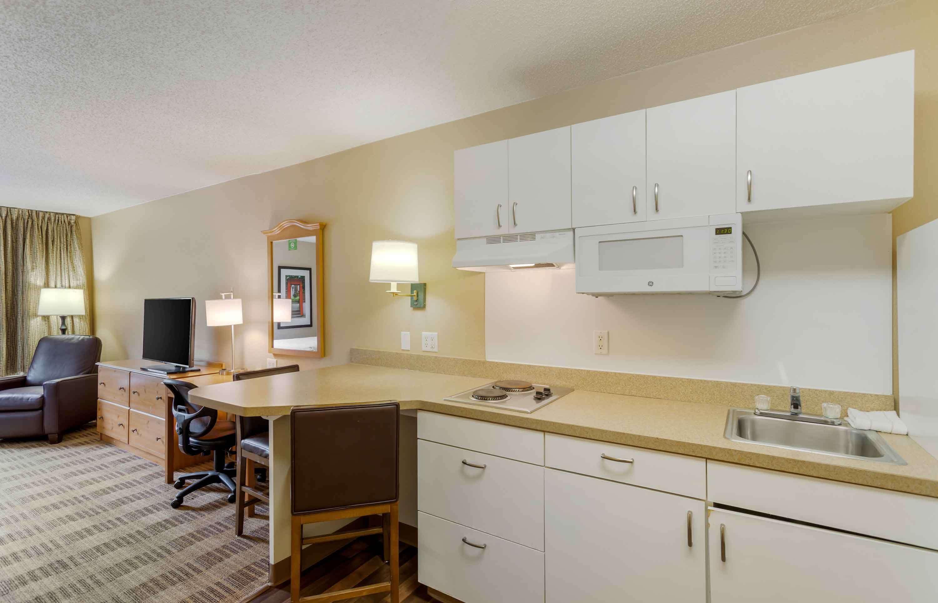 A compact hotel room with a kitchenette, a dining area, a flat-screen TV, a sofa, and a lamp.