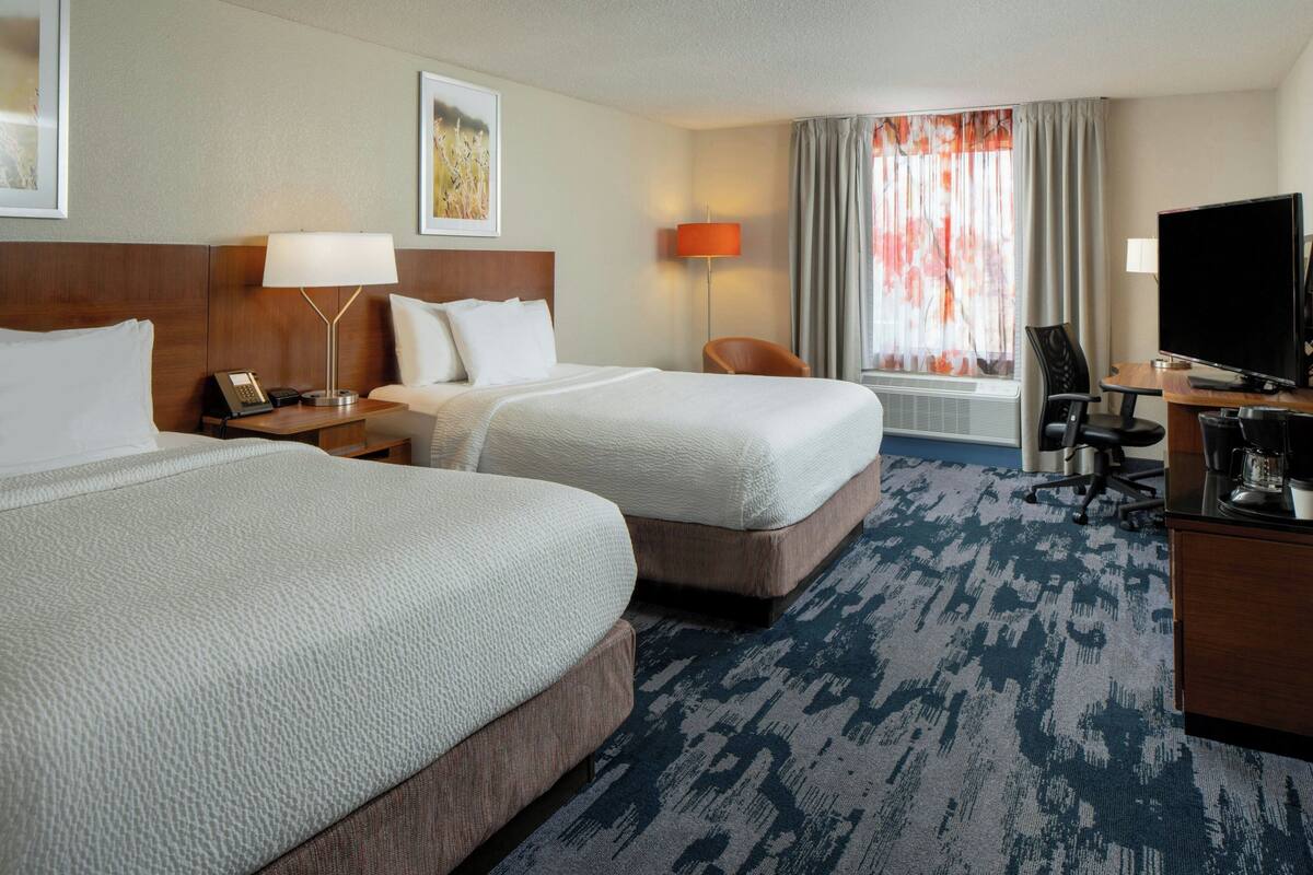 A hotel room with two beds, a desk with a computer, a chair, a TV, and a window with curtains.