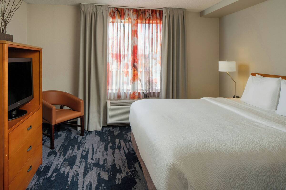 A hotel room with a bed, a desk with a television, a chair, a window with curtains, and a lamp.