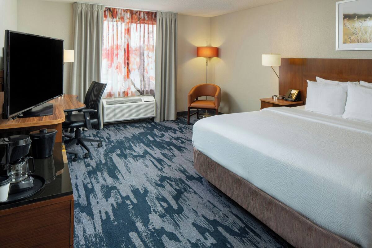 A hotel room with a large bed, a desk with a flat-screen TV, a chair, an air conditioning unit, and a lamp.