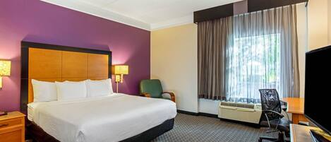 A hotel room with a large bed, a desk with a flat-screen TV, a chair, and an air conditioning unit.
