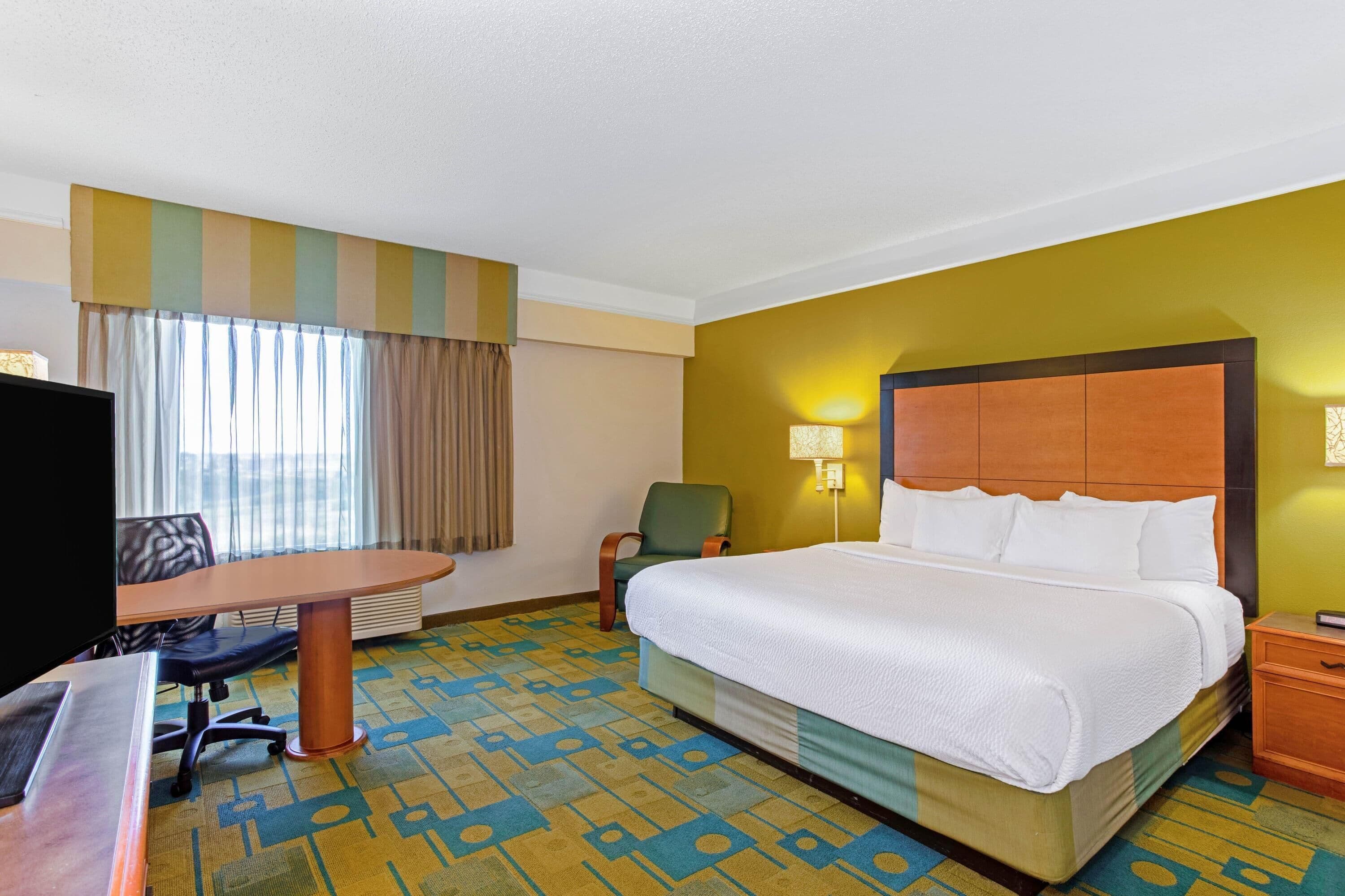 Quarto de hotel com uma cama grande, uma escrivaninha com cadeira, televisão e uma janela com cortinas.