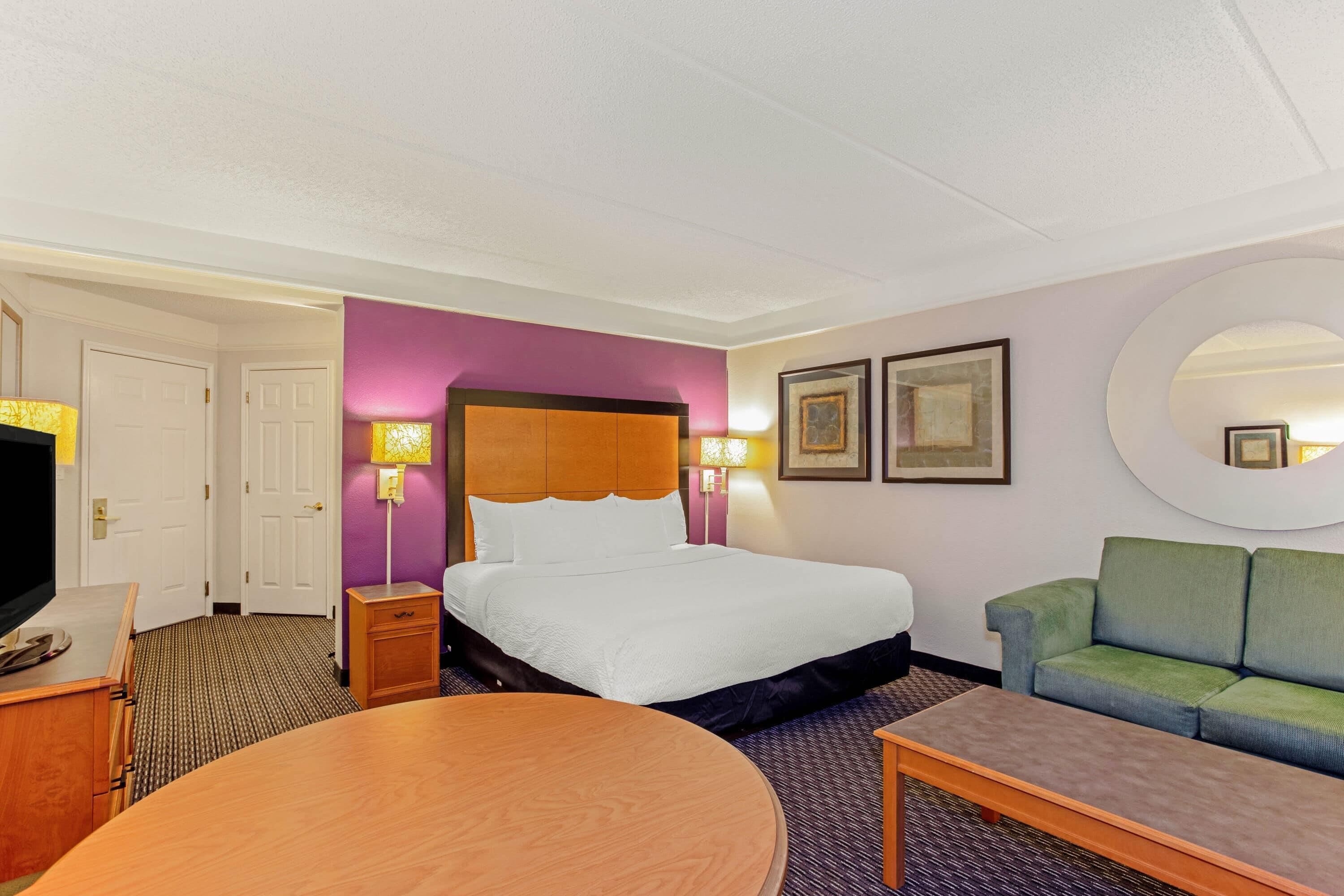 A hotel room with a bed, a green sofa, a round wooden table, a TV, and a mirror.