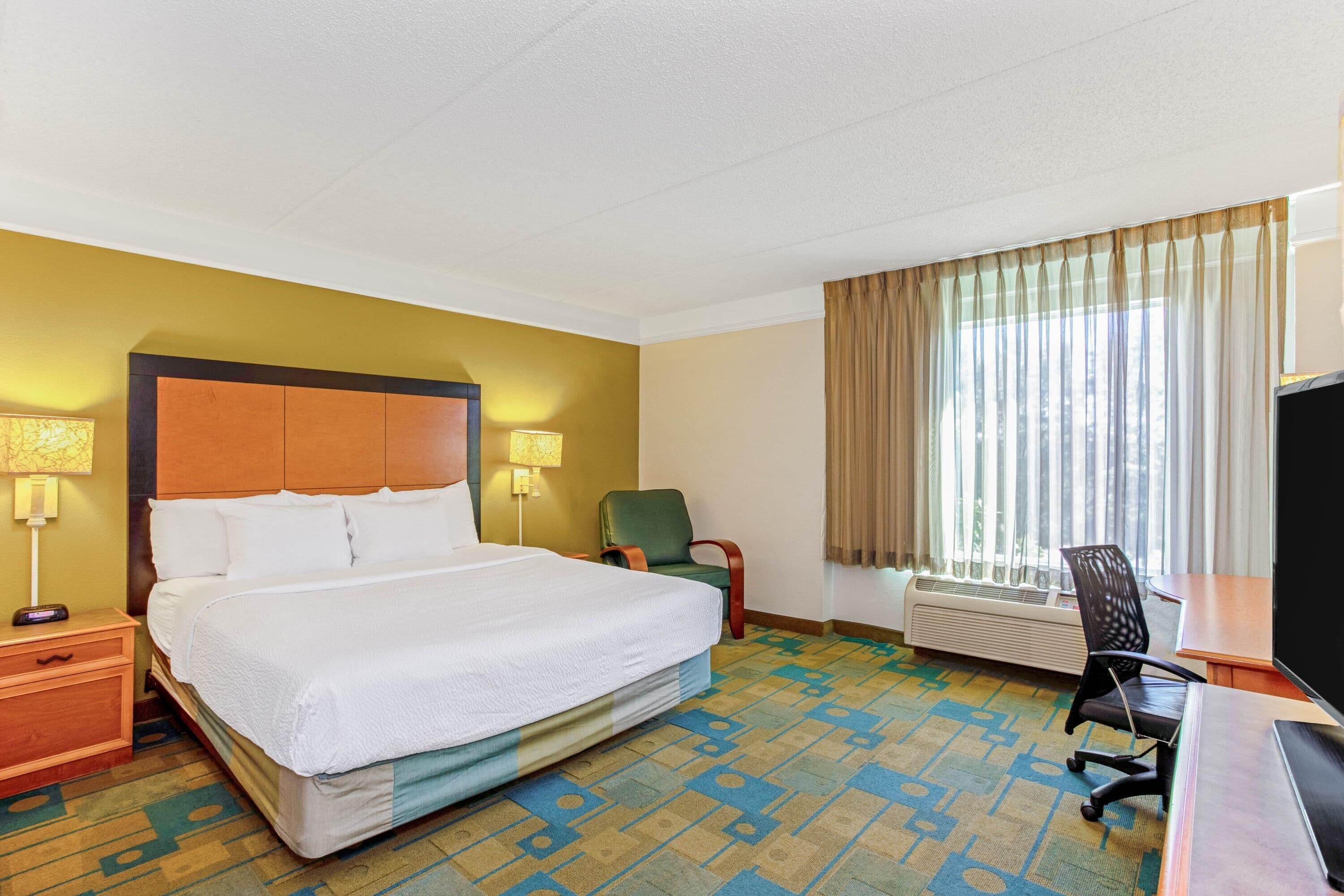 A hotel room with a bed, two bedside lamps, a desk with a chair, a window with curtains, and a wall-mounted air conditioner.