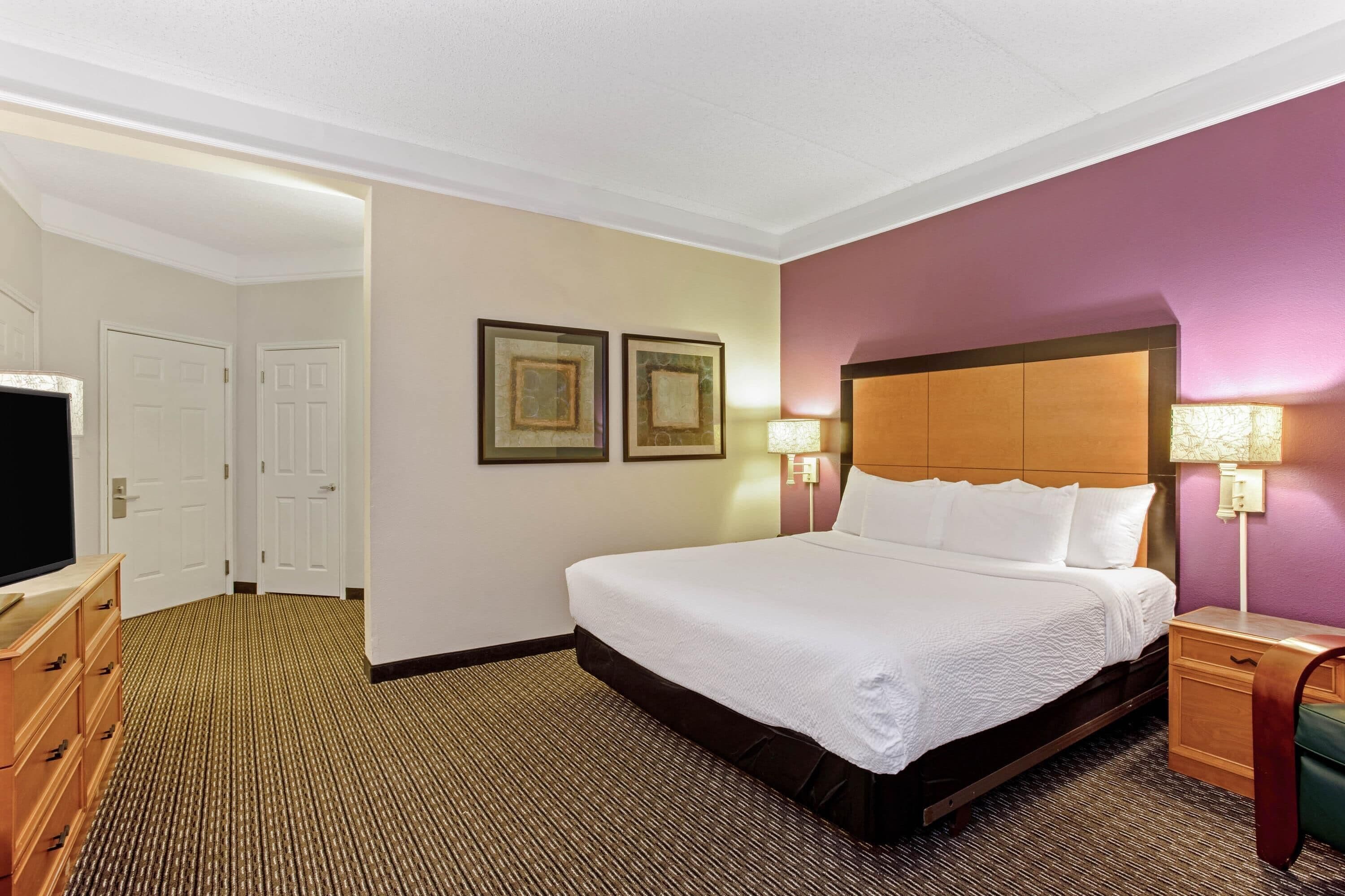 Quarto de hotel com uma cama grande, duas luminárias de cabeceira, uma cômoda de madeira e uma televisão.