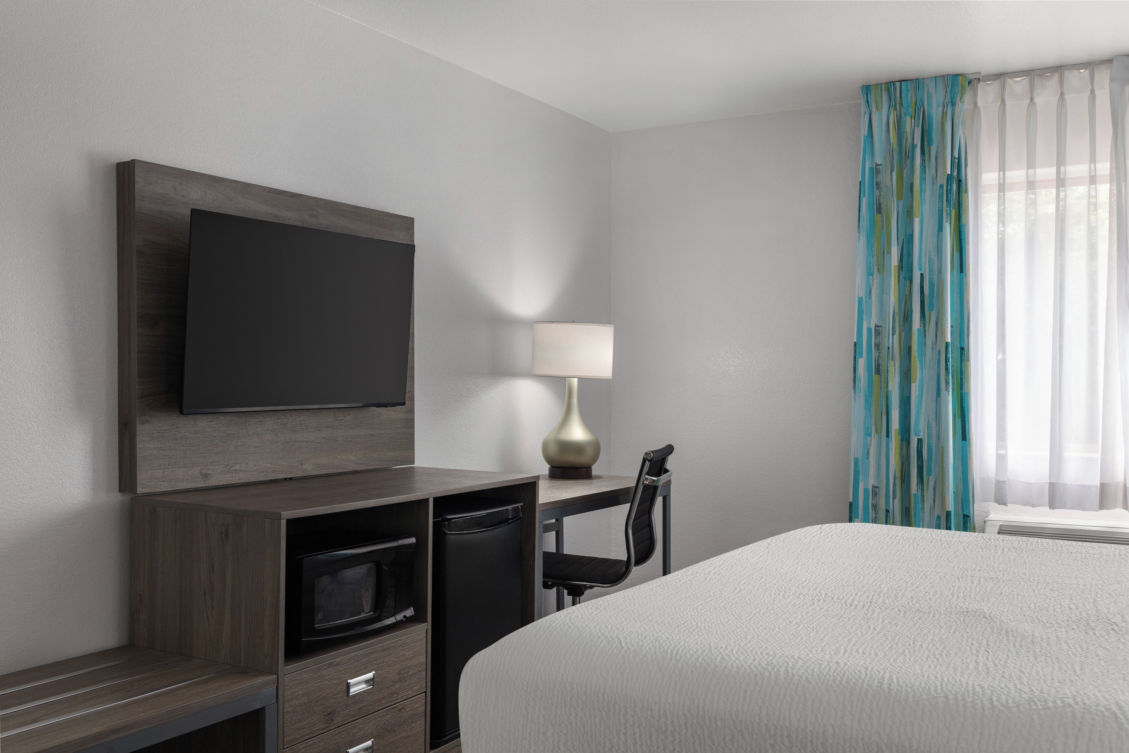 Habitación de hotel con cama, televisión montada en la pared, escritorio con silla, lámpara y ventana con cortinas.