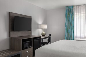 Habitación de hotel con cama, televisión montada en la pared, escritorio con silla, lámpara y ventana con cortinas.