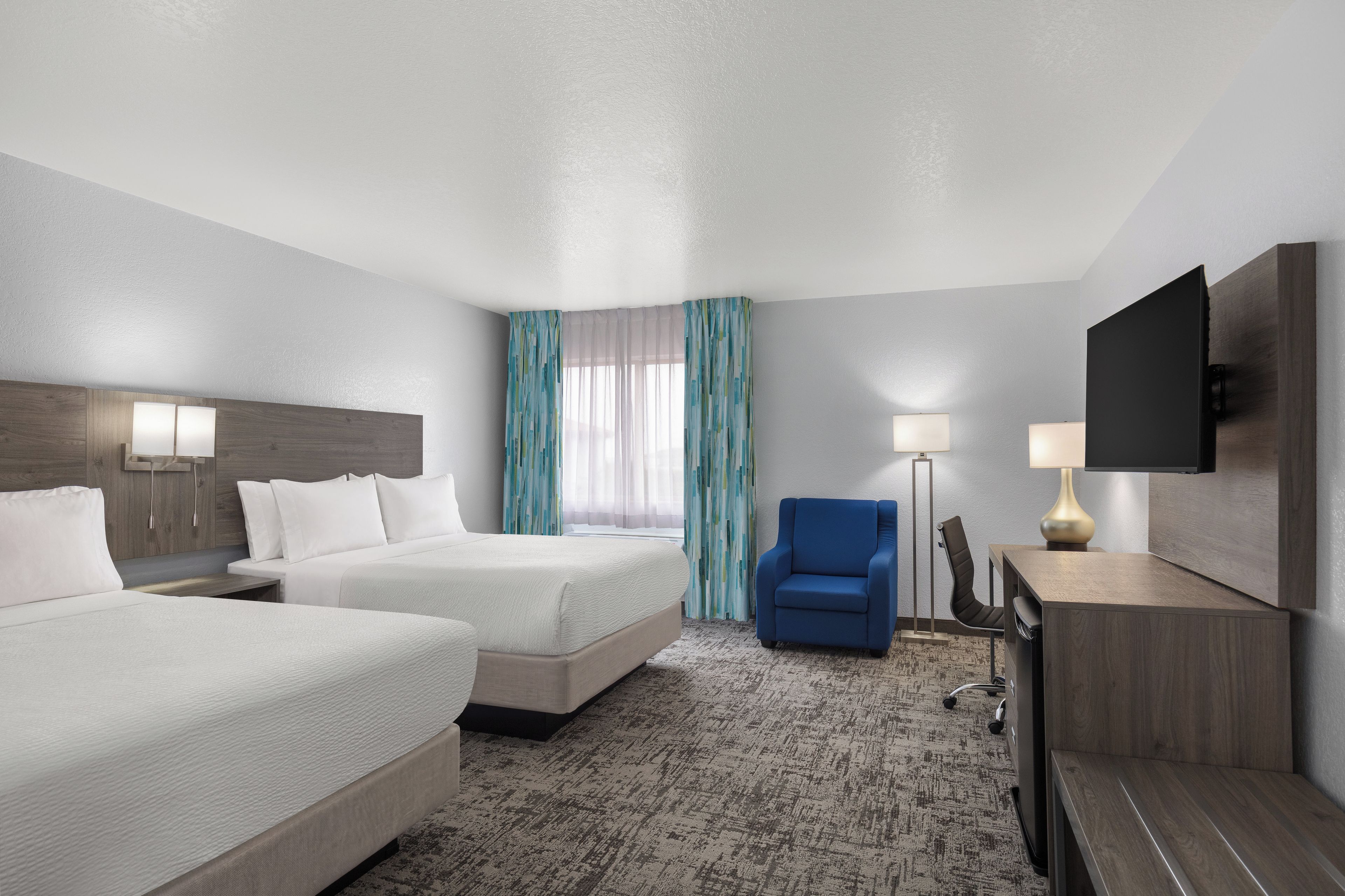 A hotel room with two beds, a desk, a chair, a TV, and a window with curtains.