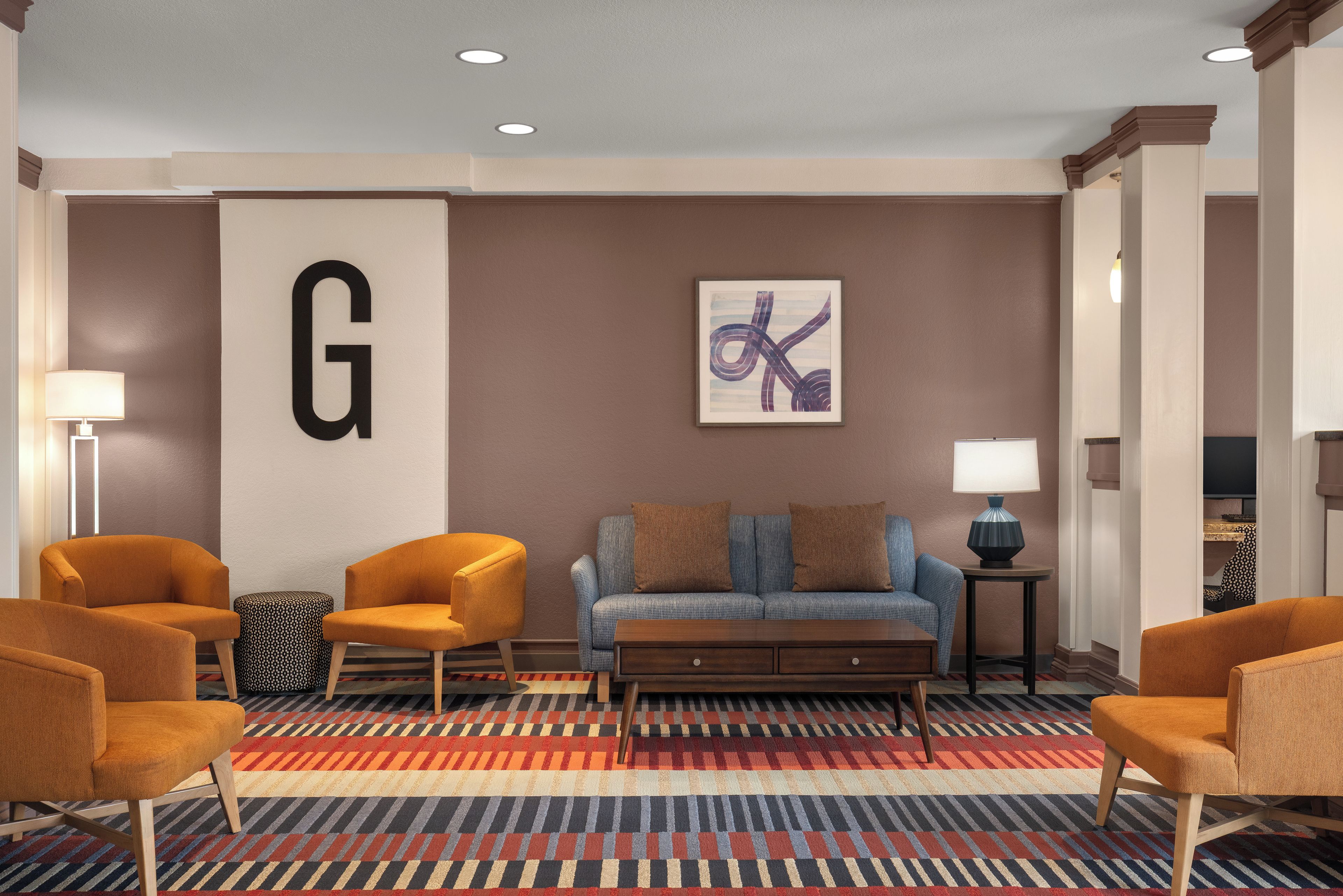A hotel lobby with a large 'G' sign, a blue sofa with brown cushions, orange armchairs, and a patterned carpet.