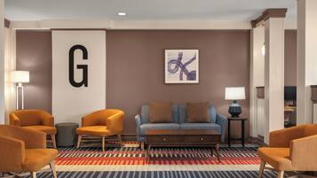 A hotel lobby with a large 'G' sign, a blue sofa with brown cushions, orange armchairs, and a patterned carpet.