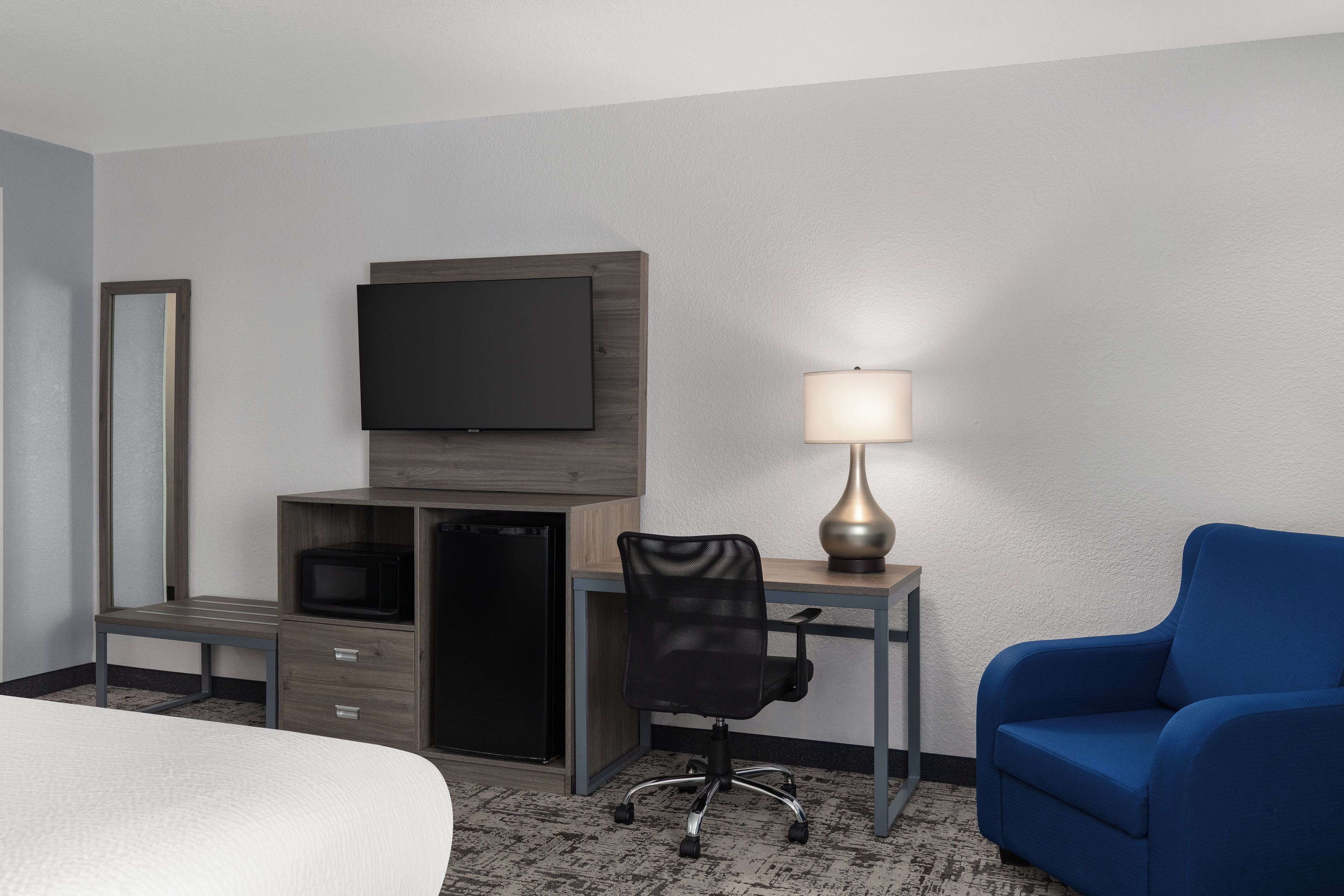 A hotel room with a bed, a desk with a television, a chair, a lamp, and a blue armchair.