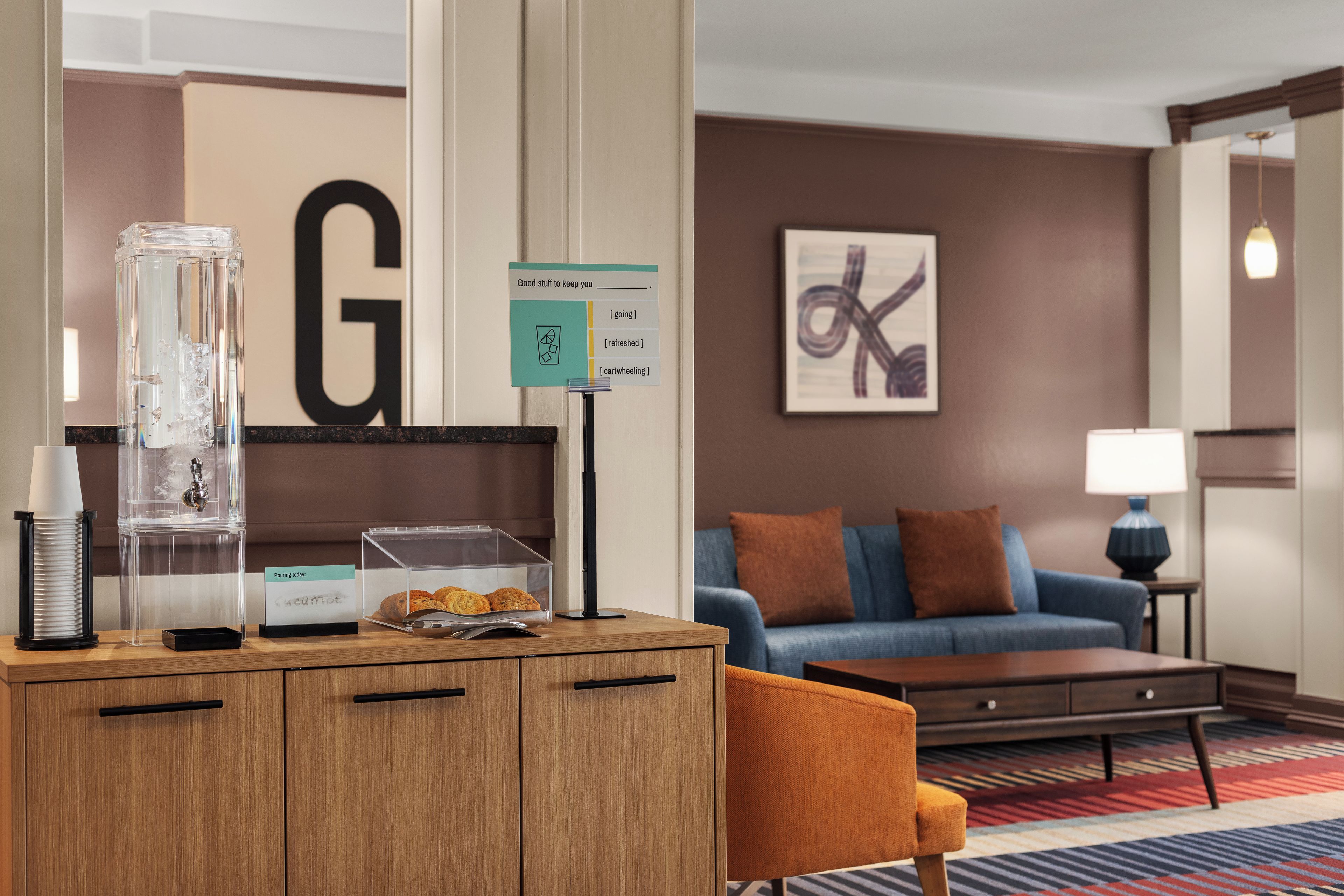 Habitación de hotel con un escritorio de madera, un recipiente de vidrio, un cartel con la letra 'G', un sofá azul y una lámpara.