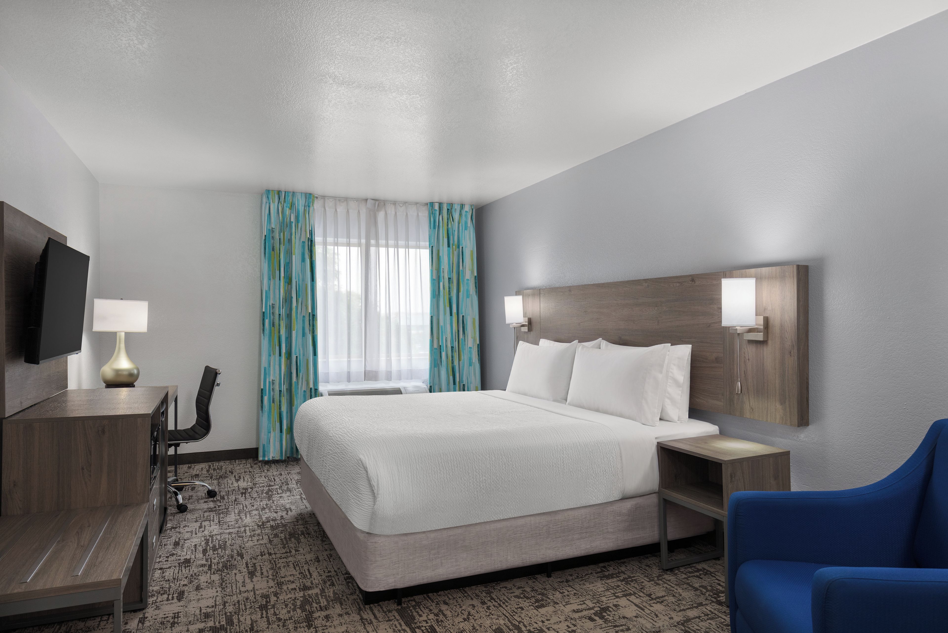 A hotel room with a bed, a desk, a chair, a TV, and a window with curtains.