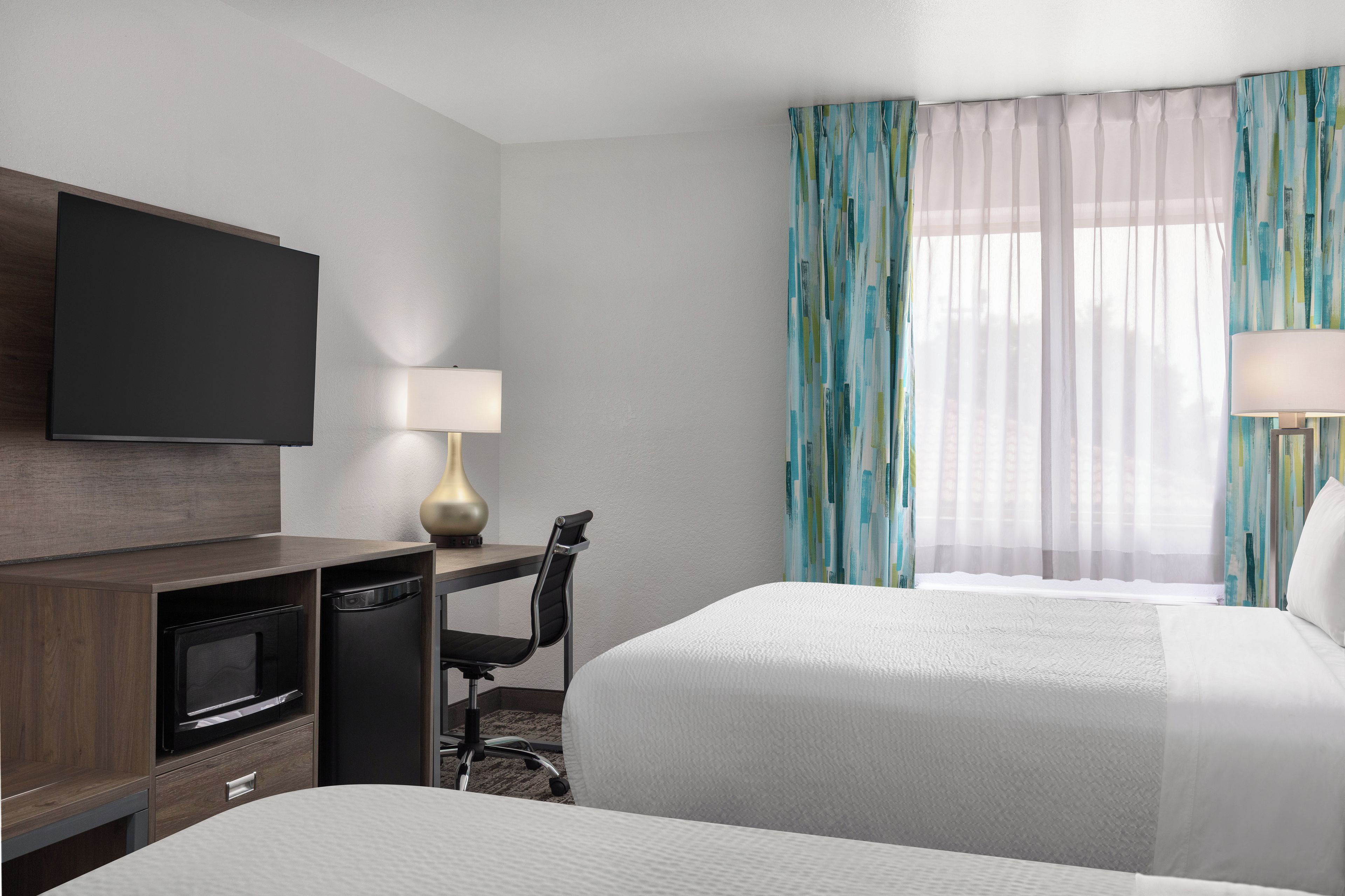 A hotel room with a bed, a desk with a lamp, a television, and a window with curtains.