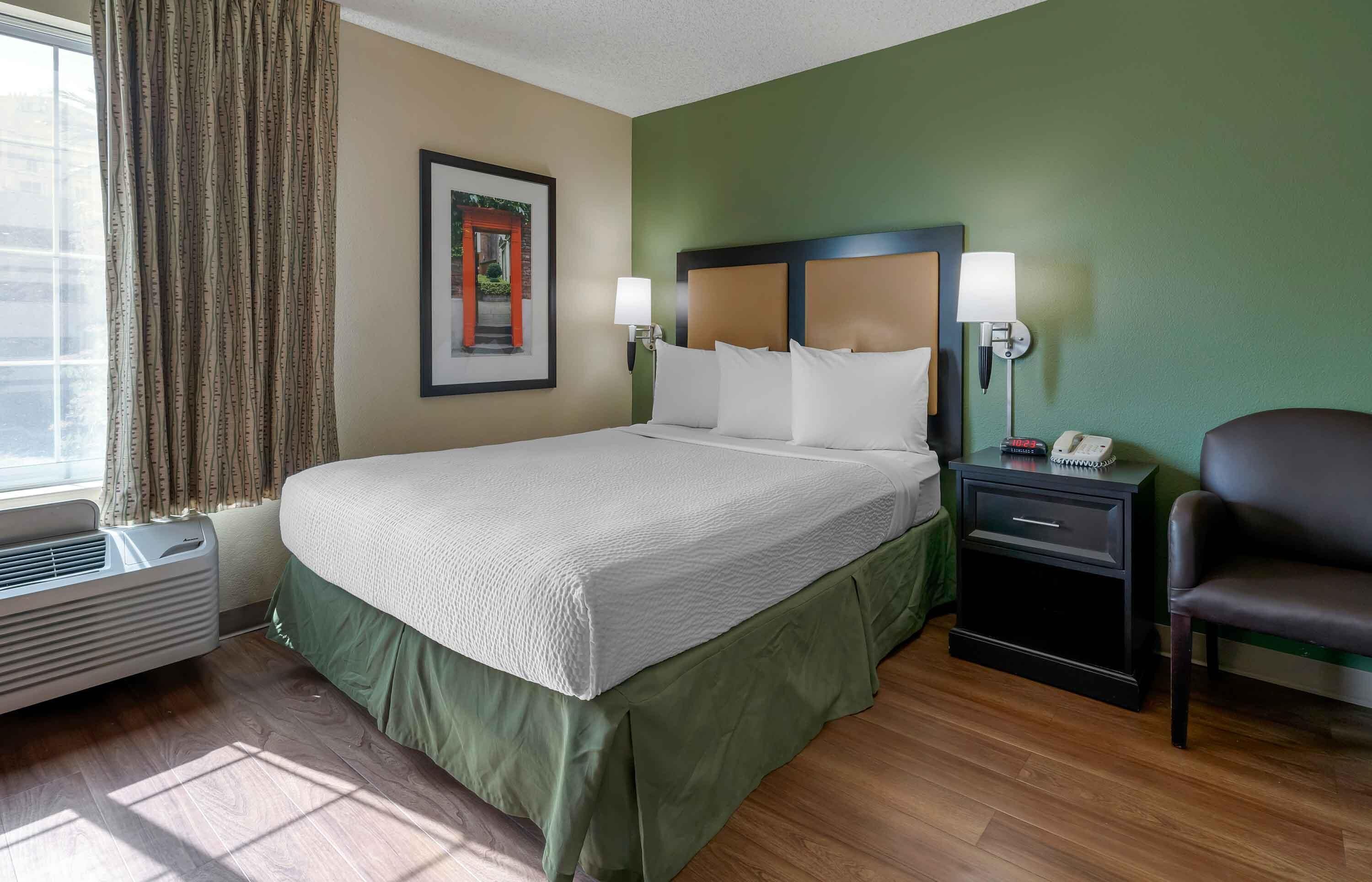 Una habitación de hotel con cama, mesita de noche, silla, un cuadro en la pared y ventana con cortinas.
