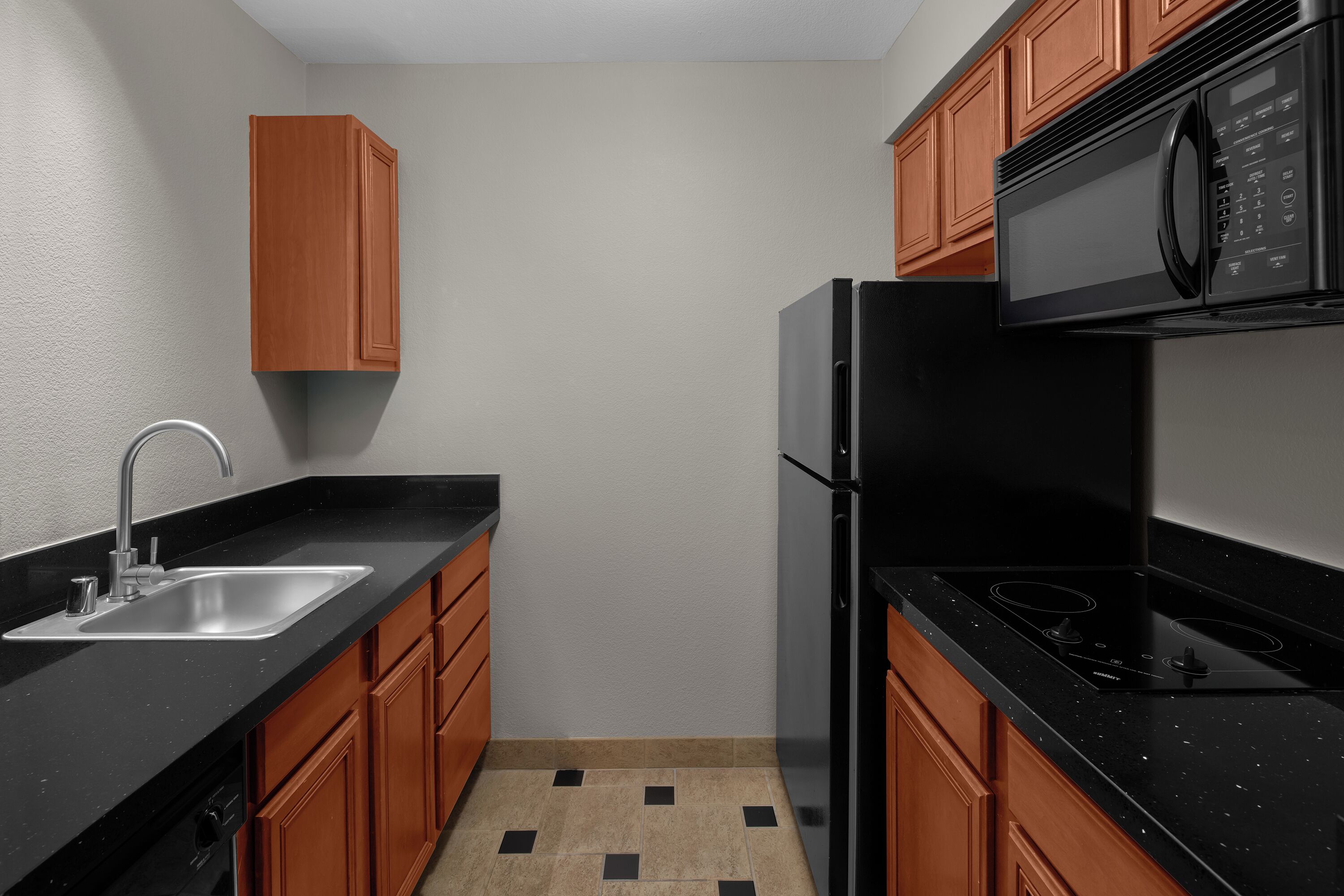 A compact kitchen with a black countertop, stainless steel sink, wooden cabinets, and a black refrigerator.