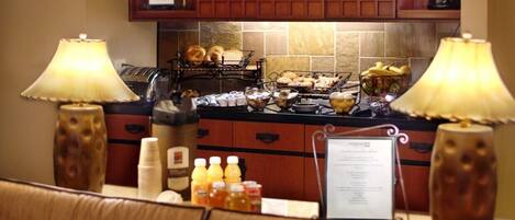 A hotel breakfast area with a counter displaying bread, pastries, and a coffee station. There are two lamps on either side of the counter, and a menu sign is visible.