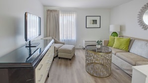 A modern living room with a flat-screen TV, a beige sofa with green pillows, a round coffee table, and a large window with sheer curtains. - Elkotel - The Newest Hotel in Elko (Elko)