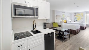 A modern kitchen with a built-in microwave, stove, and dishwasher. There is a dining area with a glass table and chairs. The room also features a bed with white bedding and a large abstract painting on the wall. - Elkotel - The Newest Hotel in Elko (Elko)
