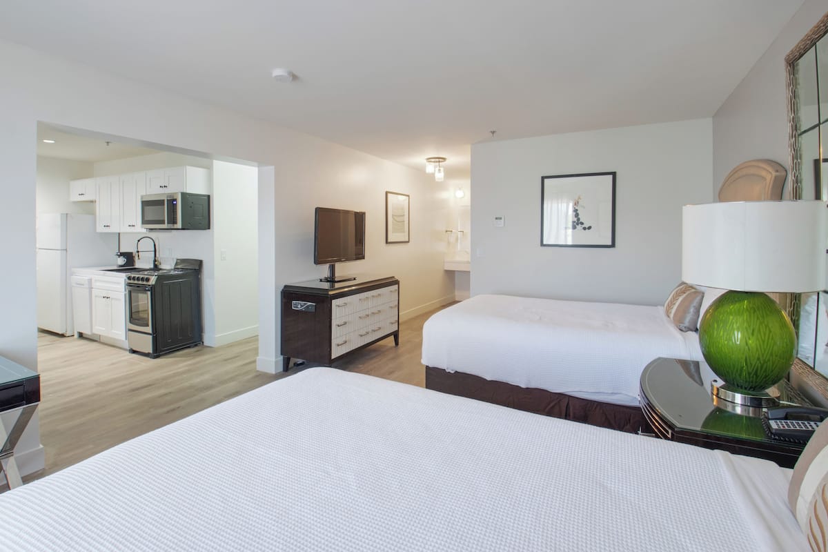 Quarto de hotel com duas camas, uma escrivaninha, cômoda, televisão e cozinha compacta.
