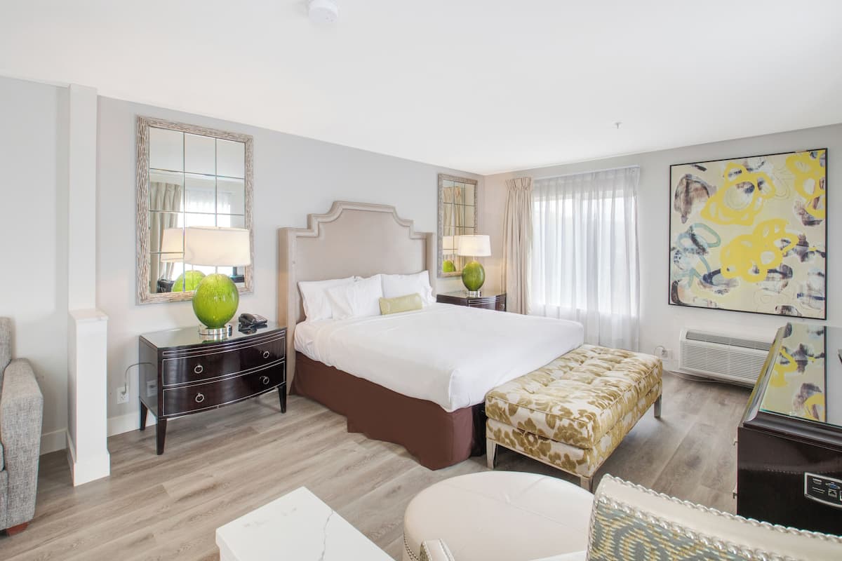 Quarto de hotel com uma cama grande, mesa de cabeceira com abajur, uma cadeira, uma pequena mesa e um quadro na parede.