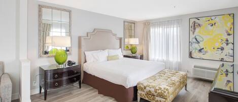 A hotel room with a large bed, a bedside table with a lamp, a chair, a small table, and a painting on the wall.