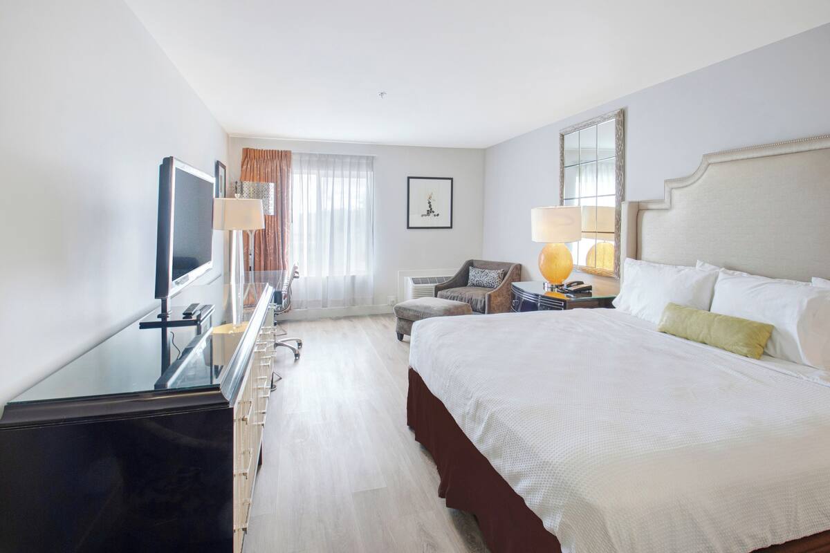 Quarto de hotel com uma cama grande, uma escrivaninha com televisão de tela plana, uma cadeira, um abajur e uma janela com cortinas.