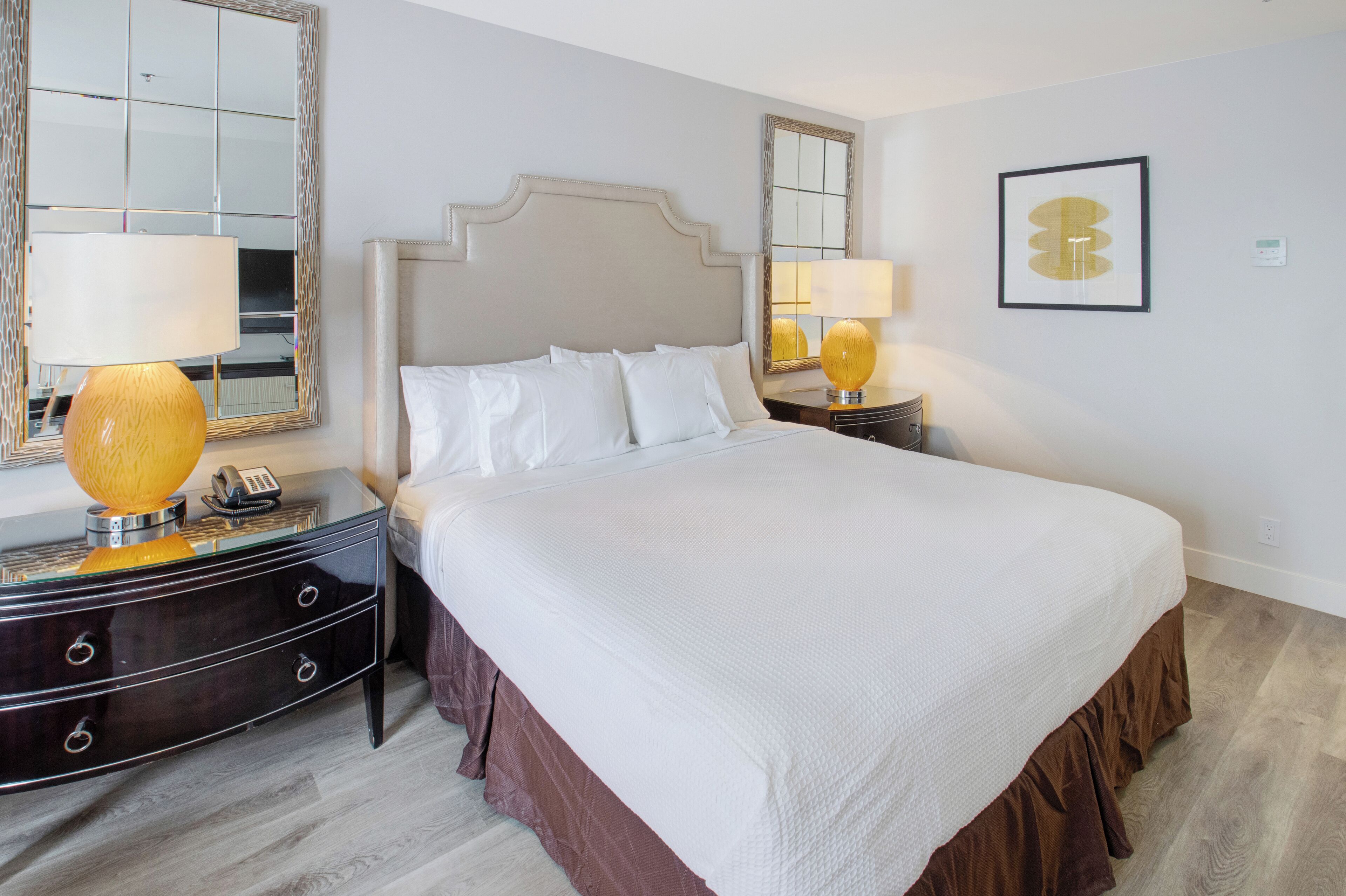 A hotel room with a large bed, two bedside tables with lamps, a mirror, and a framed artwork on the wall.
