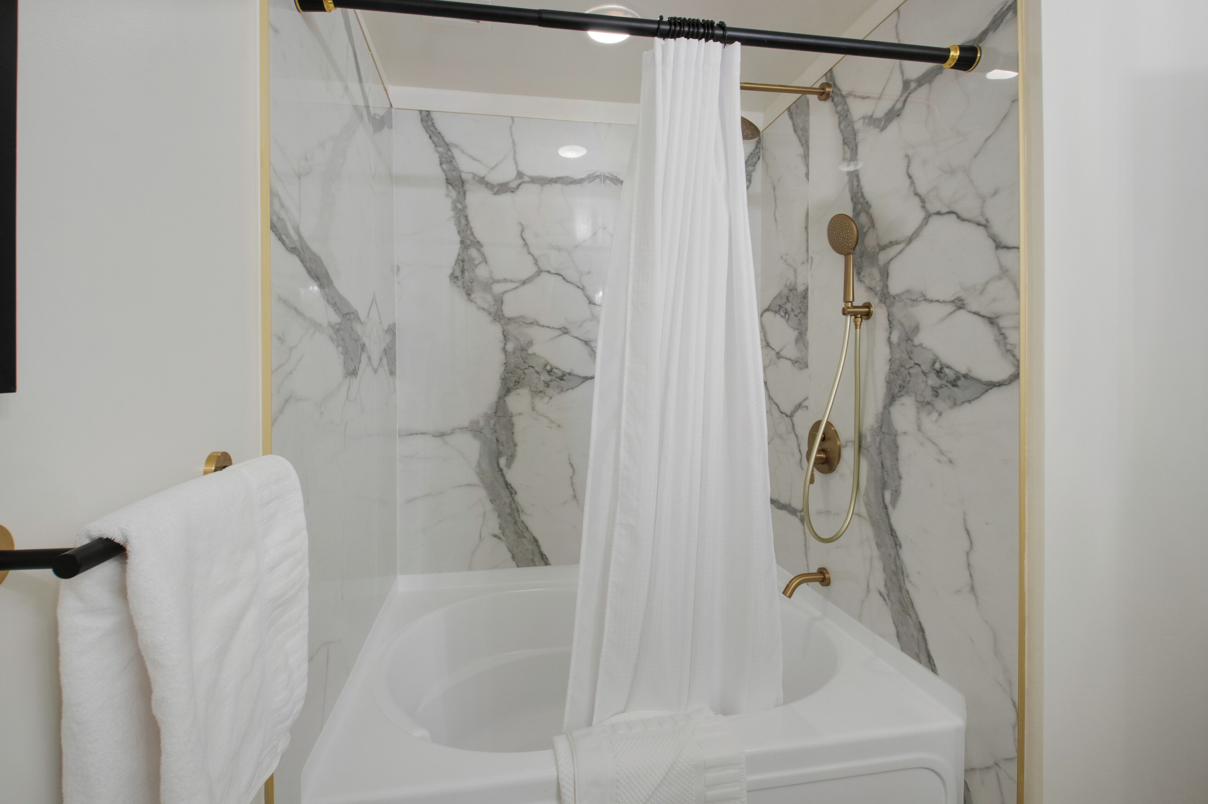 Un bagno con una parete in marmo, una vasca da bagno, una doccia con un rubinetto dorato e una tenda bianca.