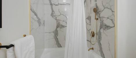 Une salle de bain avec un mur en marbre, une baignoire, une douche dotée d’un robinet doré et un rideau blanc.