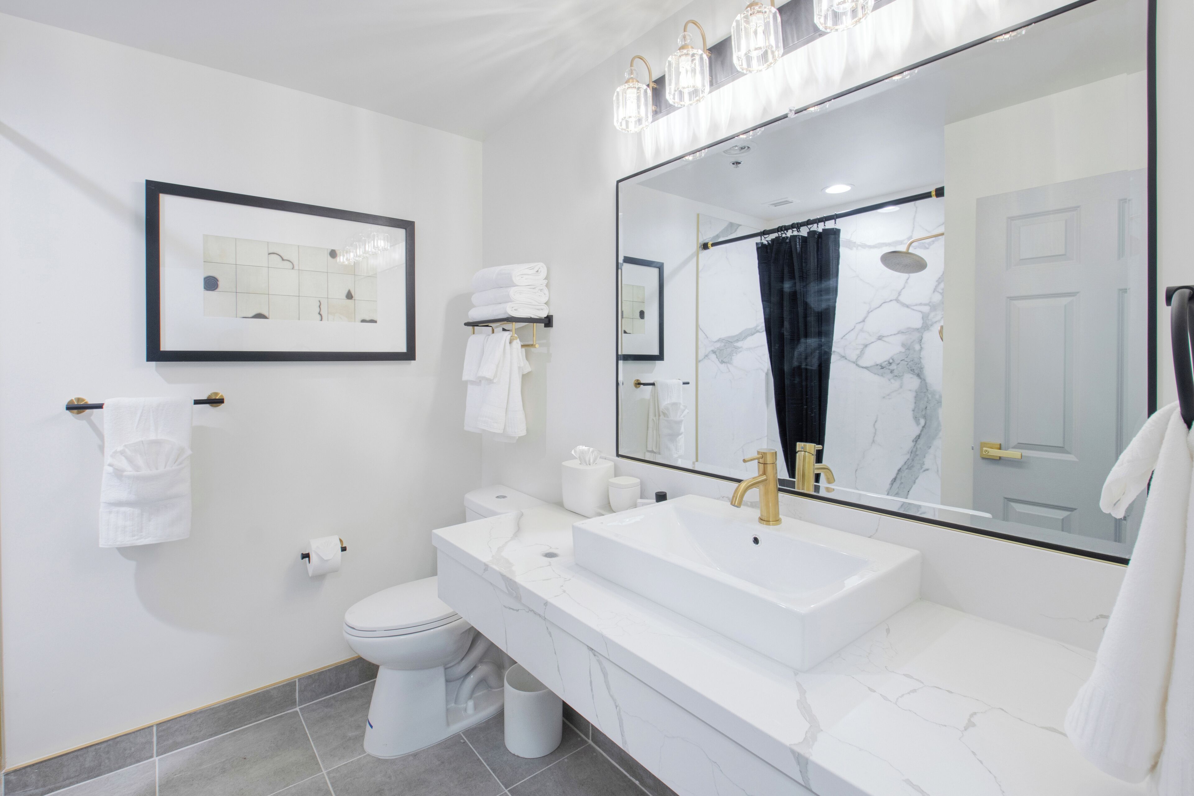 A modern bathroom with a white sink, gold faucet, and a large mirror. There is a toilet, a towel rack, and a framed picture on the wall.