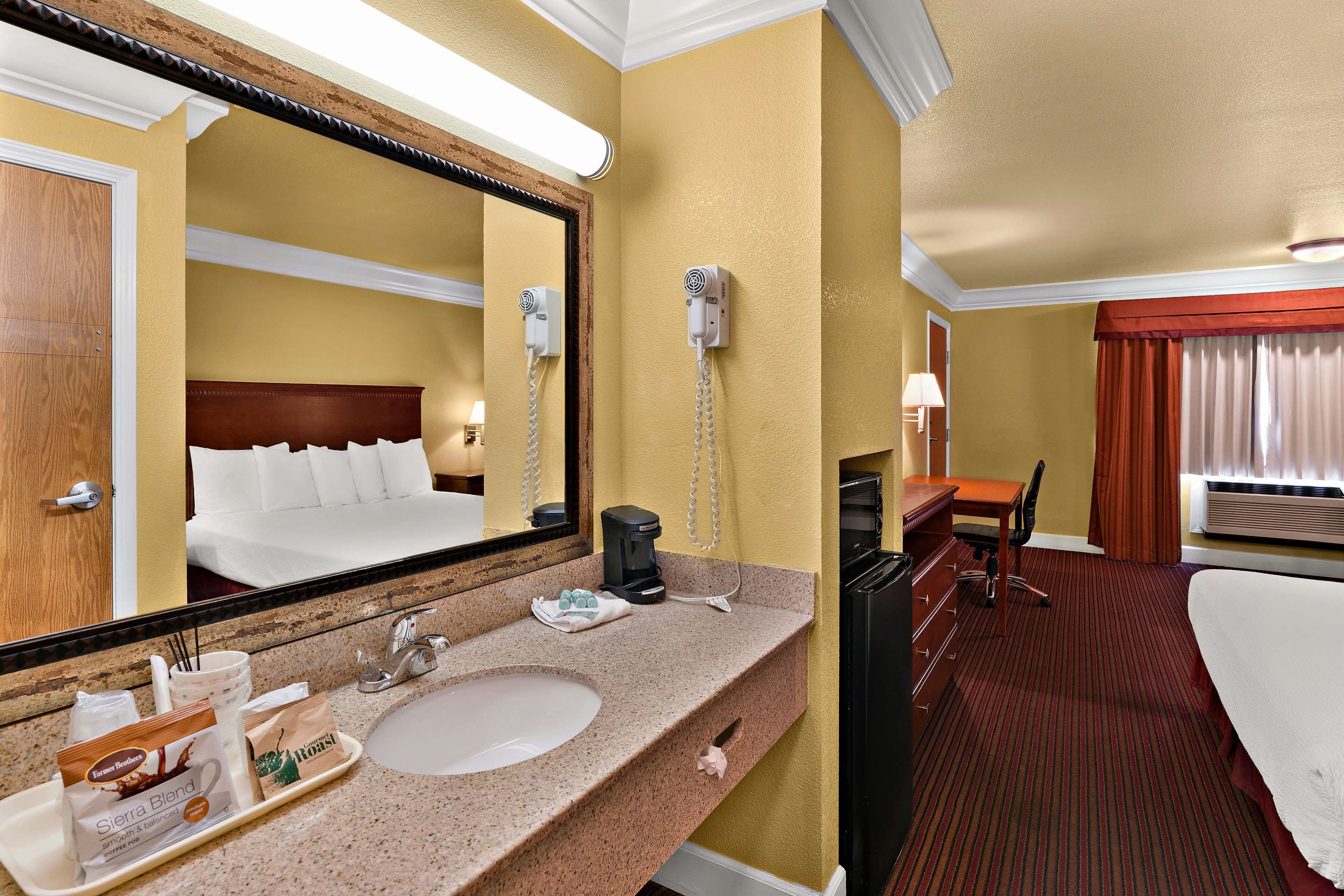 A hotel room with a large bed, a desk, a chair, a desk lamp, a hairdryer, a coffee maker, and a tray with breakfast items.