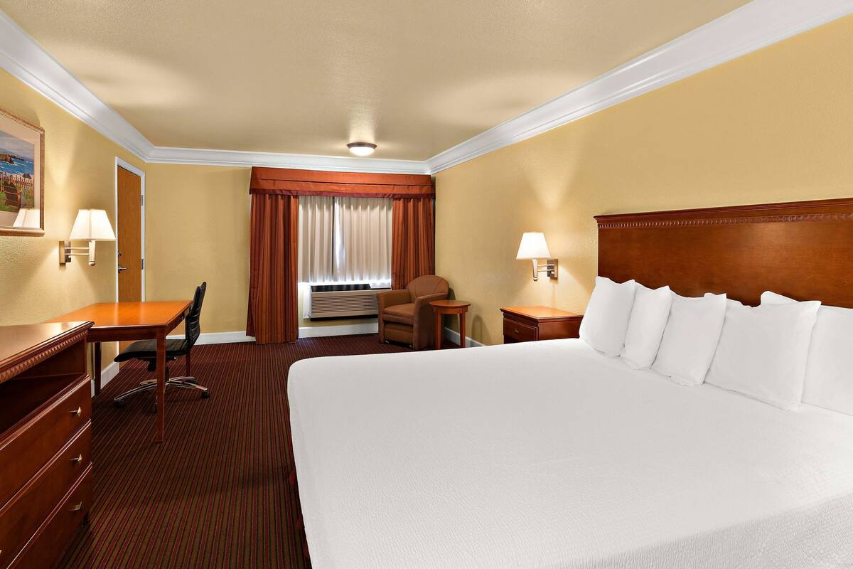 Quarto de hotel com uma cama grande, uma escrivaninha, uma cadeira, uma mesa pequena e um abajur.