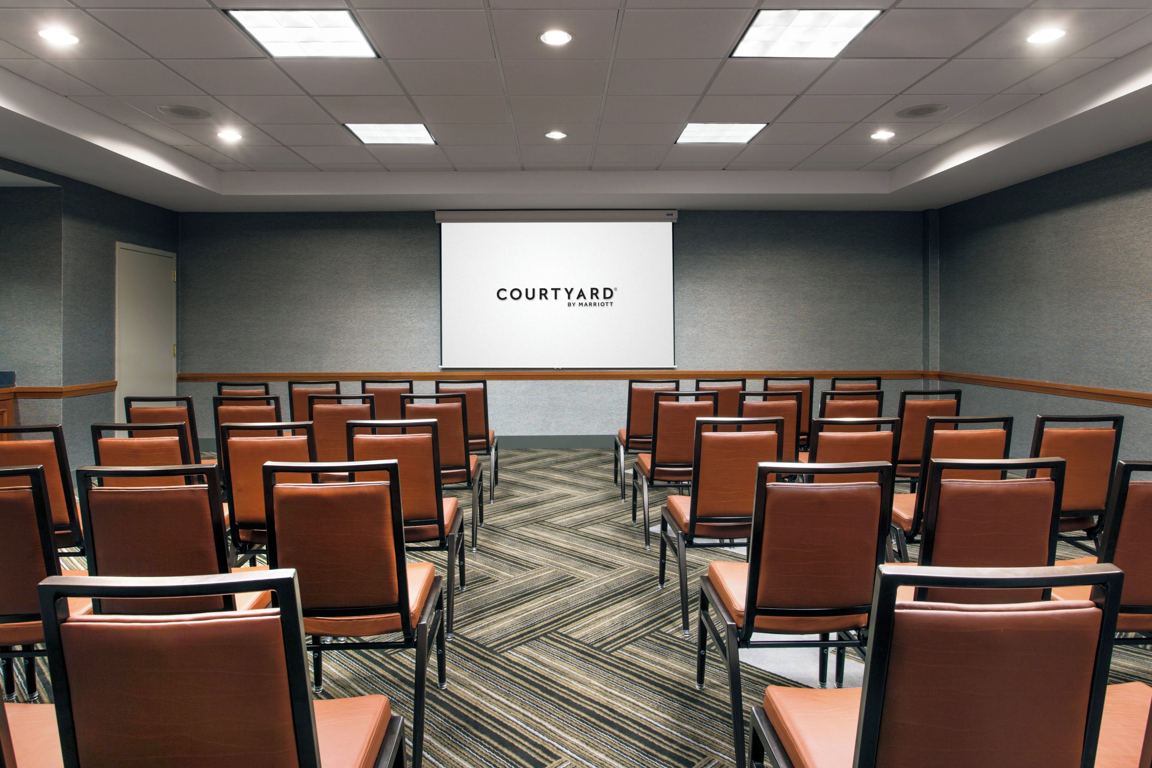 Un salón de conferencias con filas de sillas, piso alfombrado y una pantalla mostrando 'COURTYARD BY MARRIOTT'.