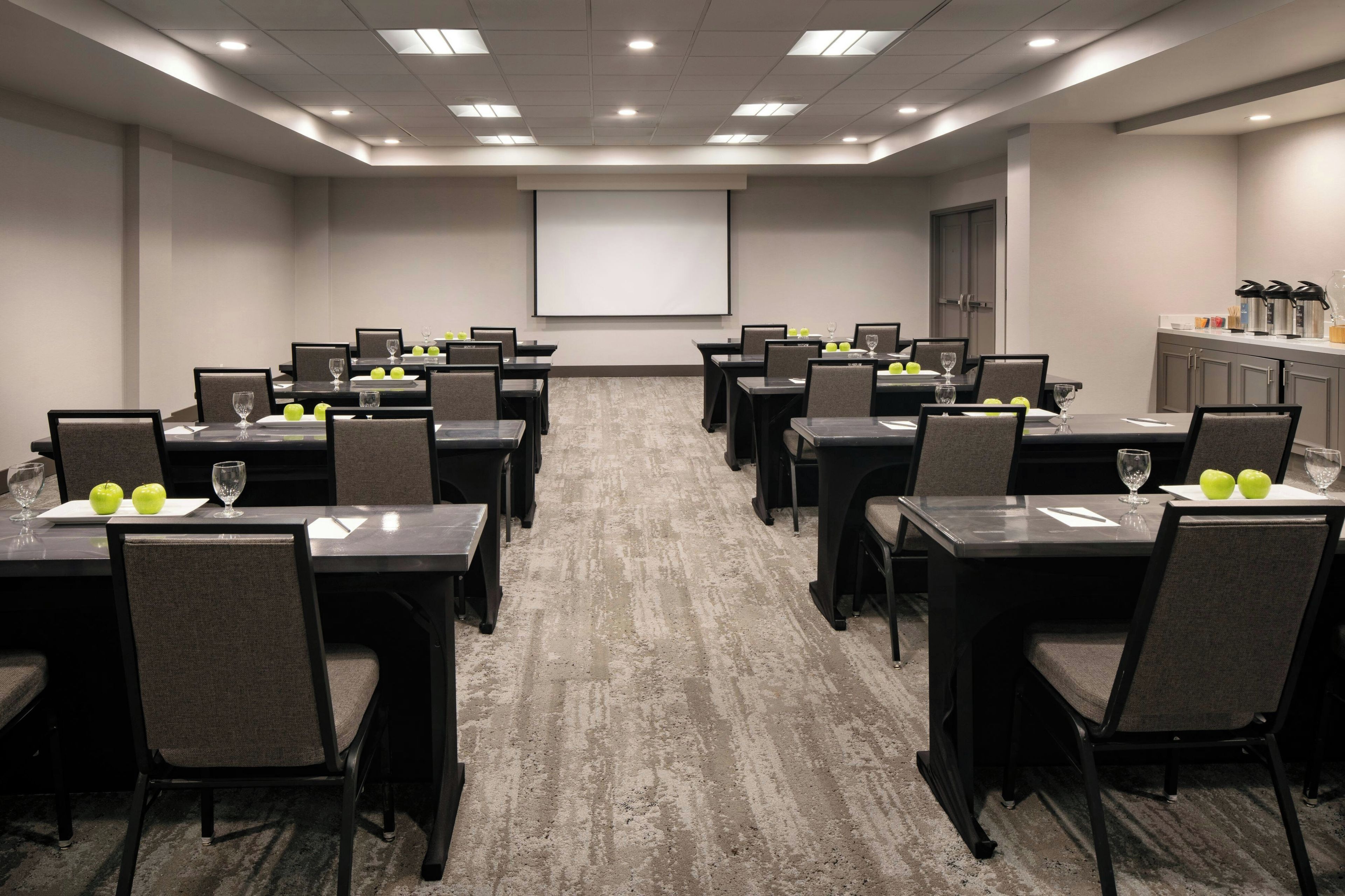 Un salón de conferencias con varias mesas, sillas, una pantalla de proyector y una zona de kitchenette.