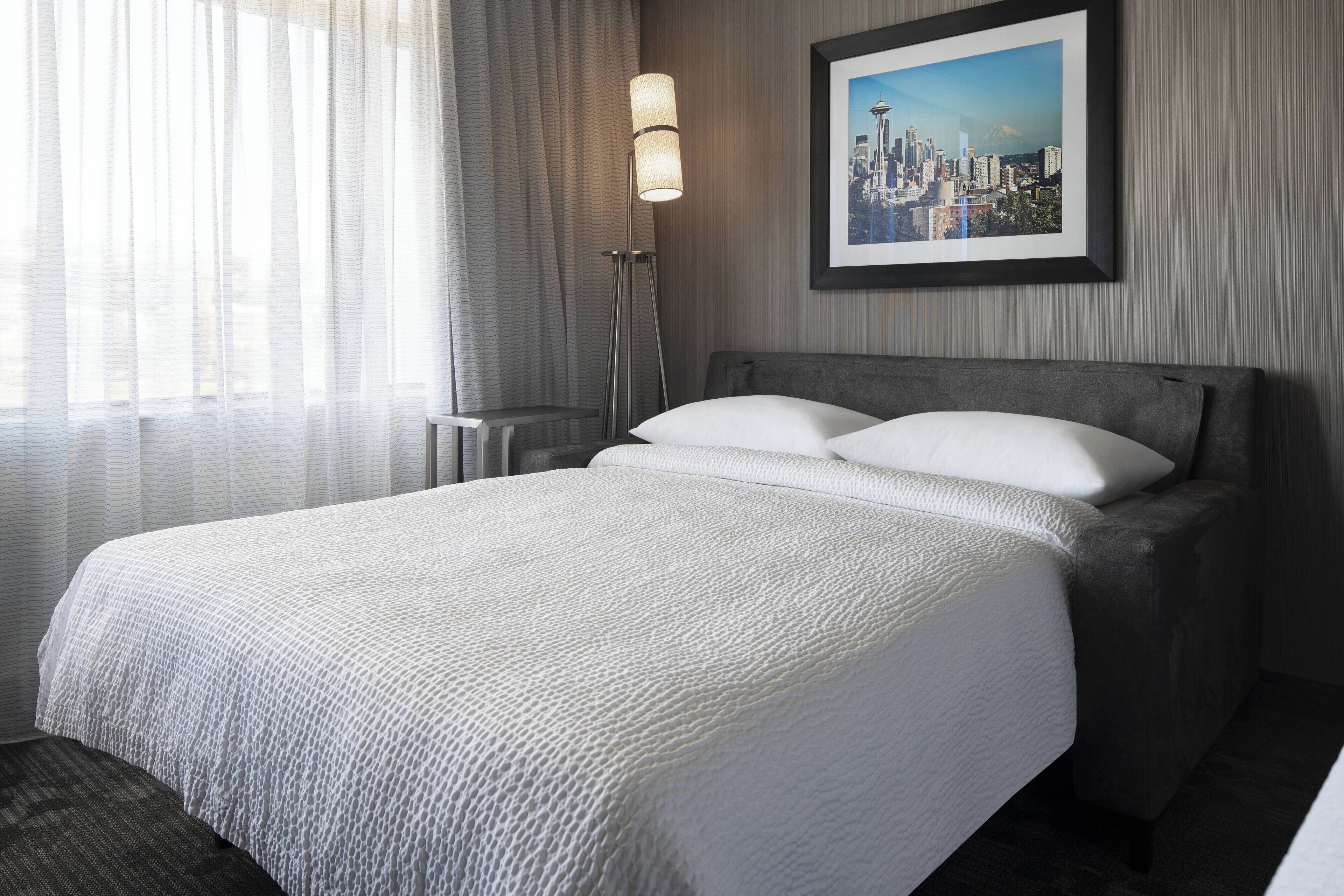 A hotel room with a large bed, a framed cityscape picture on the wall, and a floor lamp.