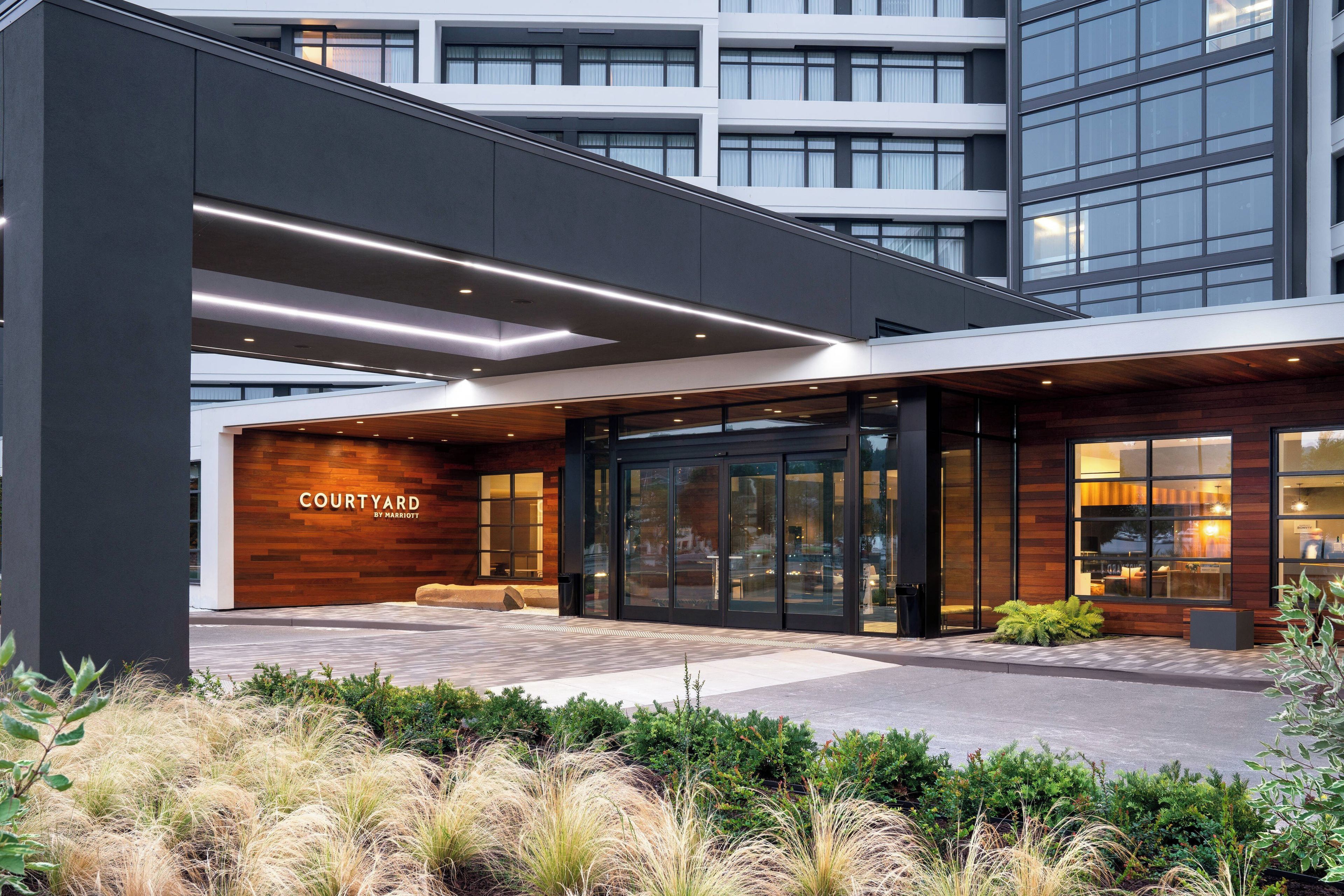 A modern hotel entrance with a sign reading 'Courtyard by Marriott,' featuring a glass facade, wooden accents, and a landscaped area in front.