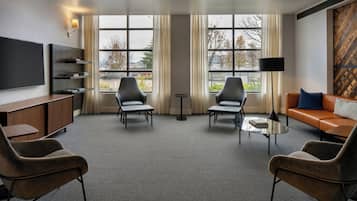 A modern hotel room with a flat-screen TV, a sofa, two armchairs, a coffee table, and a bookshelf.