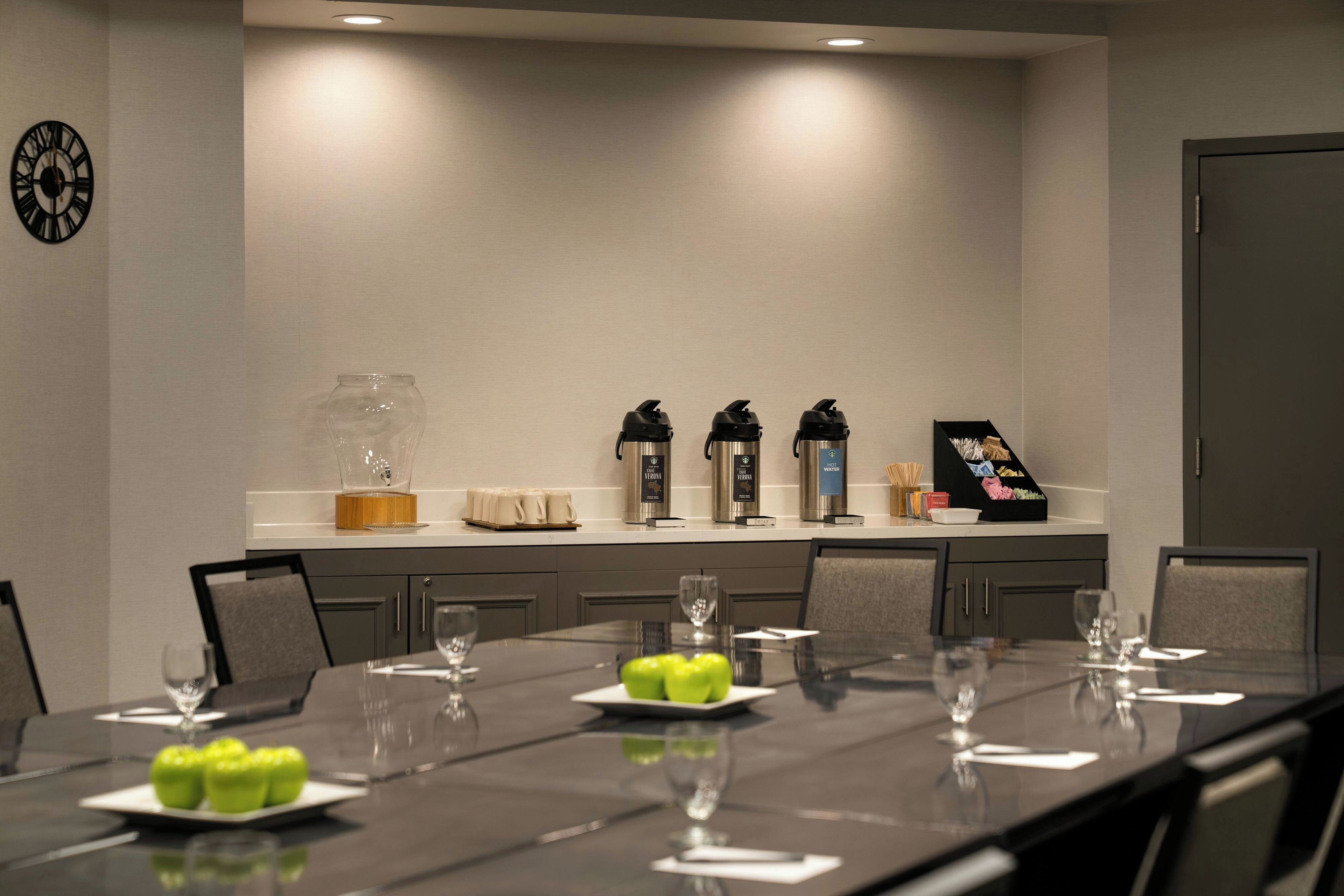 A conference room with a long table set for meetings, equipped with coffee makers, a water dispenser, and a clock on the wall.