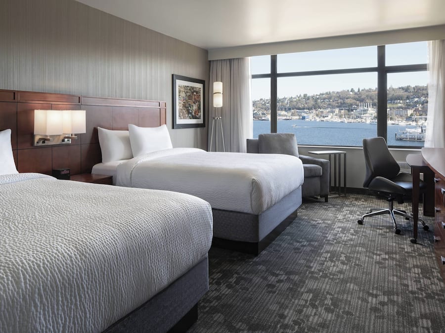 Ein Hotelzimmer mit zwei Betten, einem Schreibtisch, einem Stuhl, einer Lampe und einem großen Fenster mit Blick auf die Stadt.