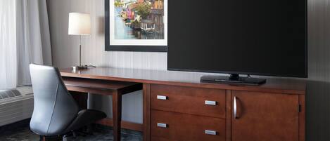 Quarto de hotel com escrivaninha de madeira, uma cadeira, televisão de tela plana, quadro pendurado na parede e luminária.
