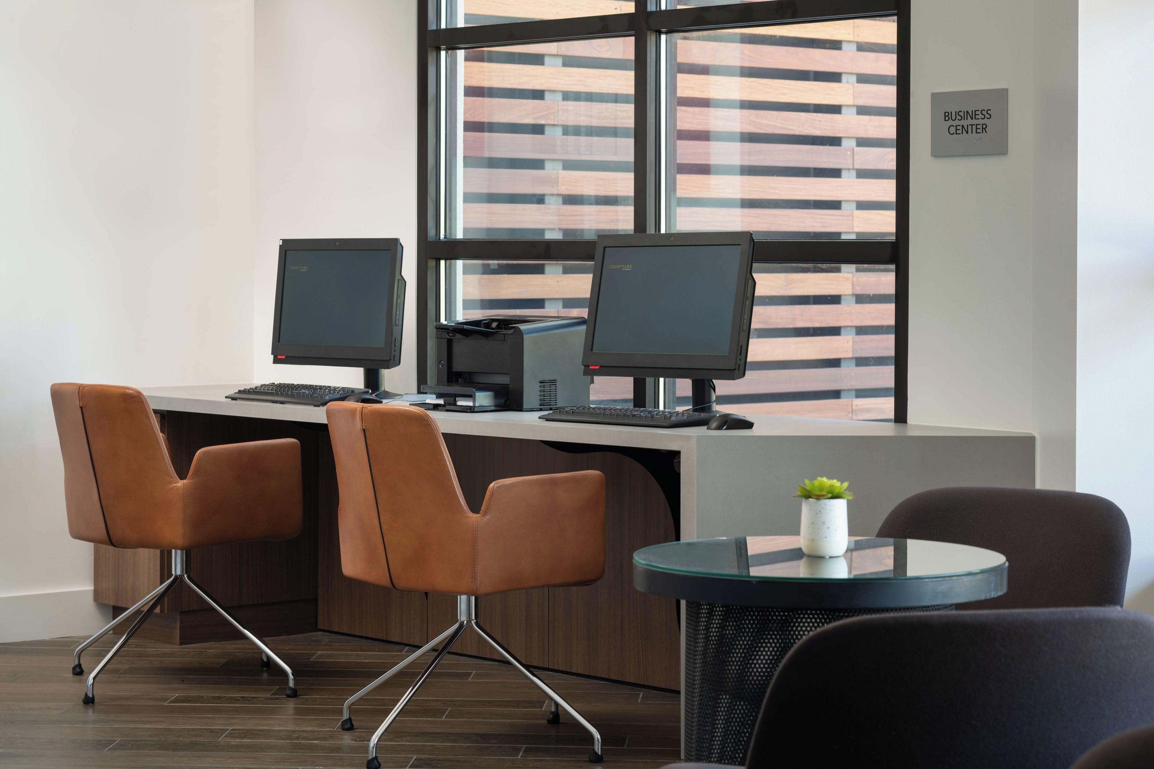 A modern office space with two desks, each with a computer monitor, keyboard, and mouse. There are three chairs, one at each desk and one on a small round table. The room has a large window with blinds, and a sign indicating a business center.