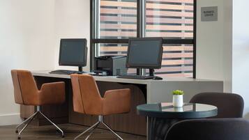 A modern office space with two desks, each with a computer monitor, keyboard, and mouse. There are three chairs, one at each desk and one on a small round table. The room has a large window with blinds, and a sign indicating a business center.