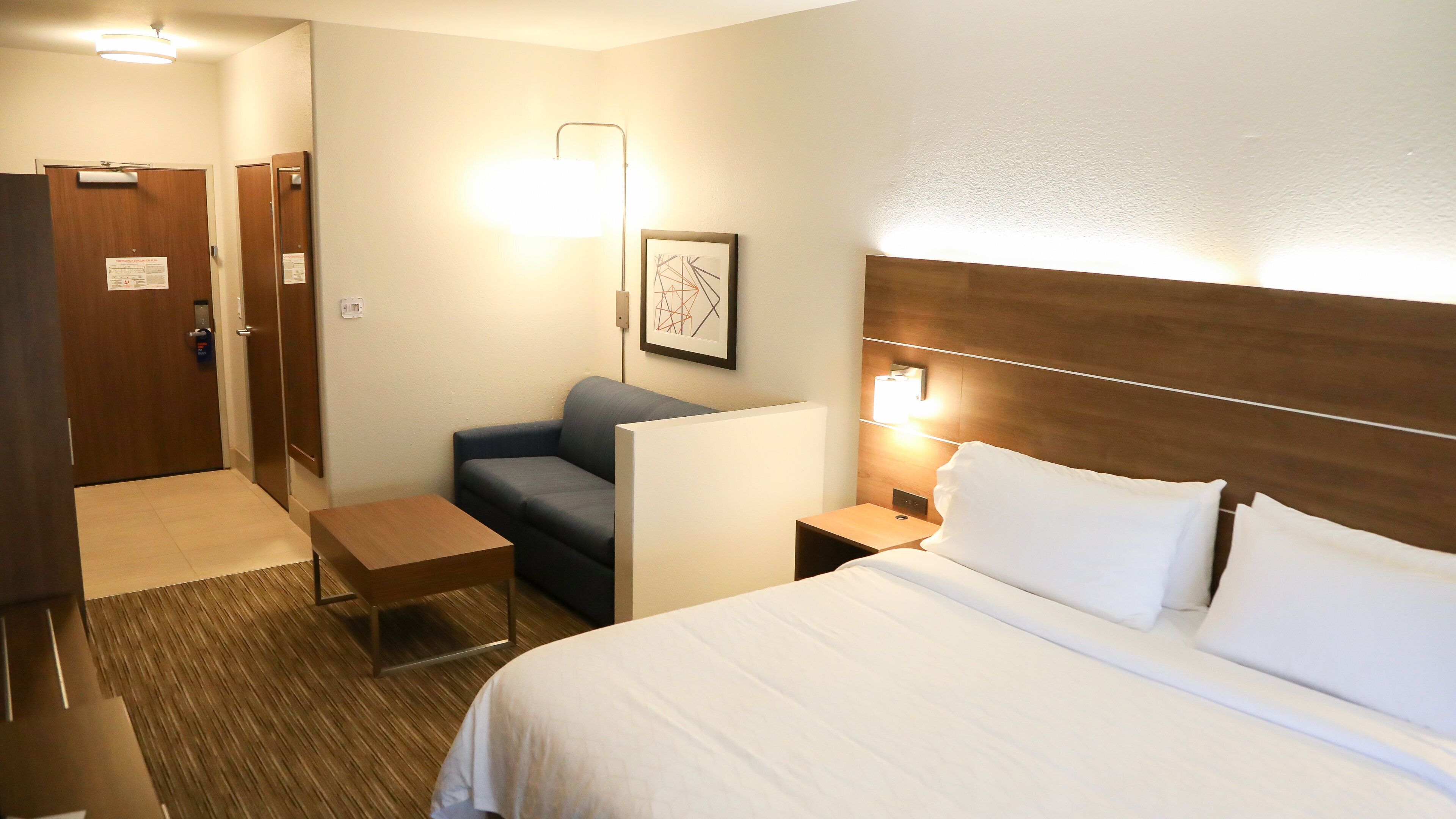 A hotel room with a bed, a desk, a chair, a sofa, and a wall-mounted light.