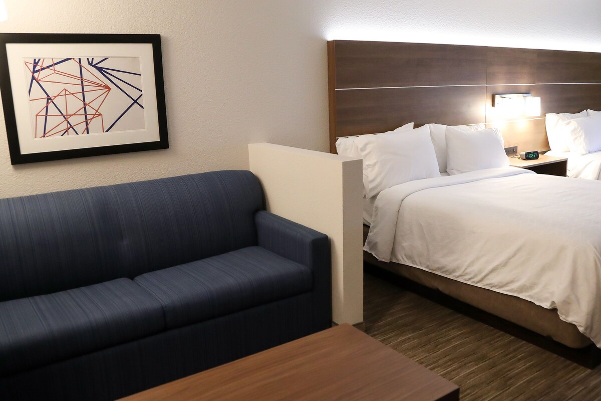 Ein Hotelzimmer mit Bett, Sofa, einem gerahmten abstrakten Kunstwerk und einem Nachttisch mit Lampe.