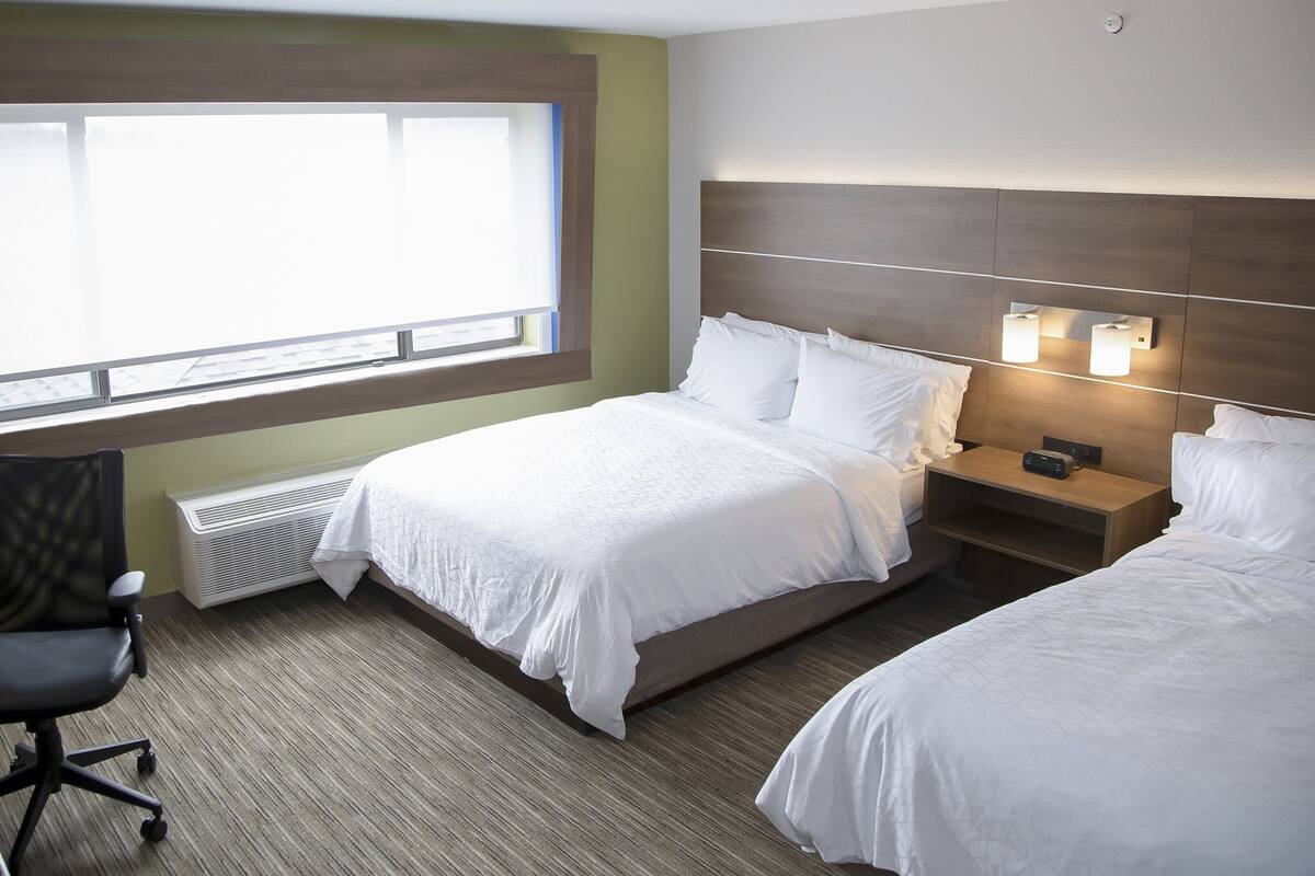 A hotel room with a large window, a desk with a chair, two beds, and a nightstand with a lamp.