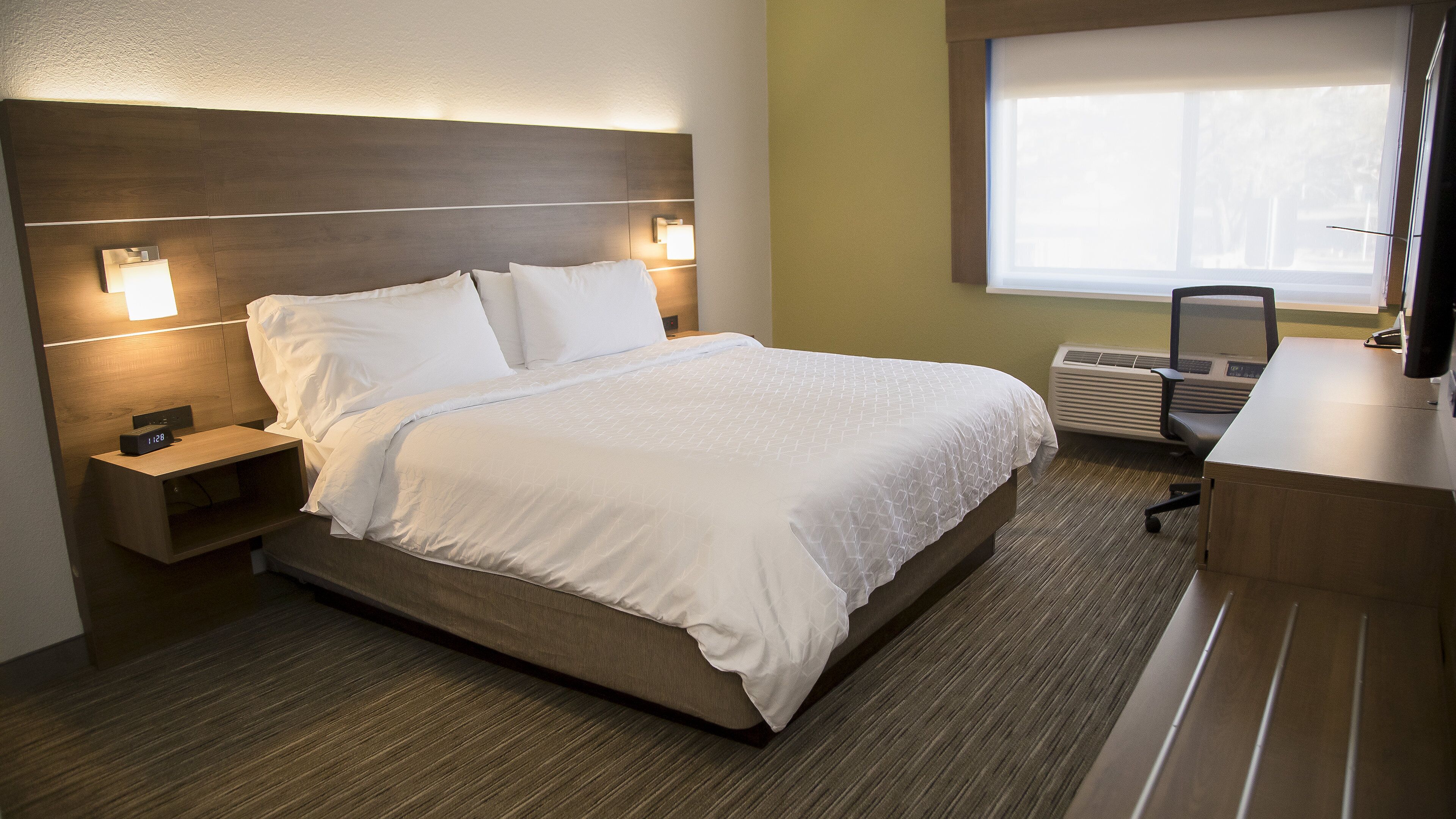 A hotel room with a bed, bedside table, lamp, and a desk with a chair.